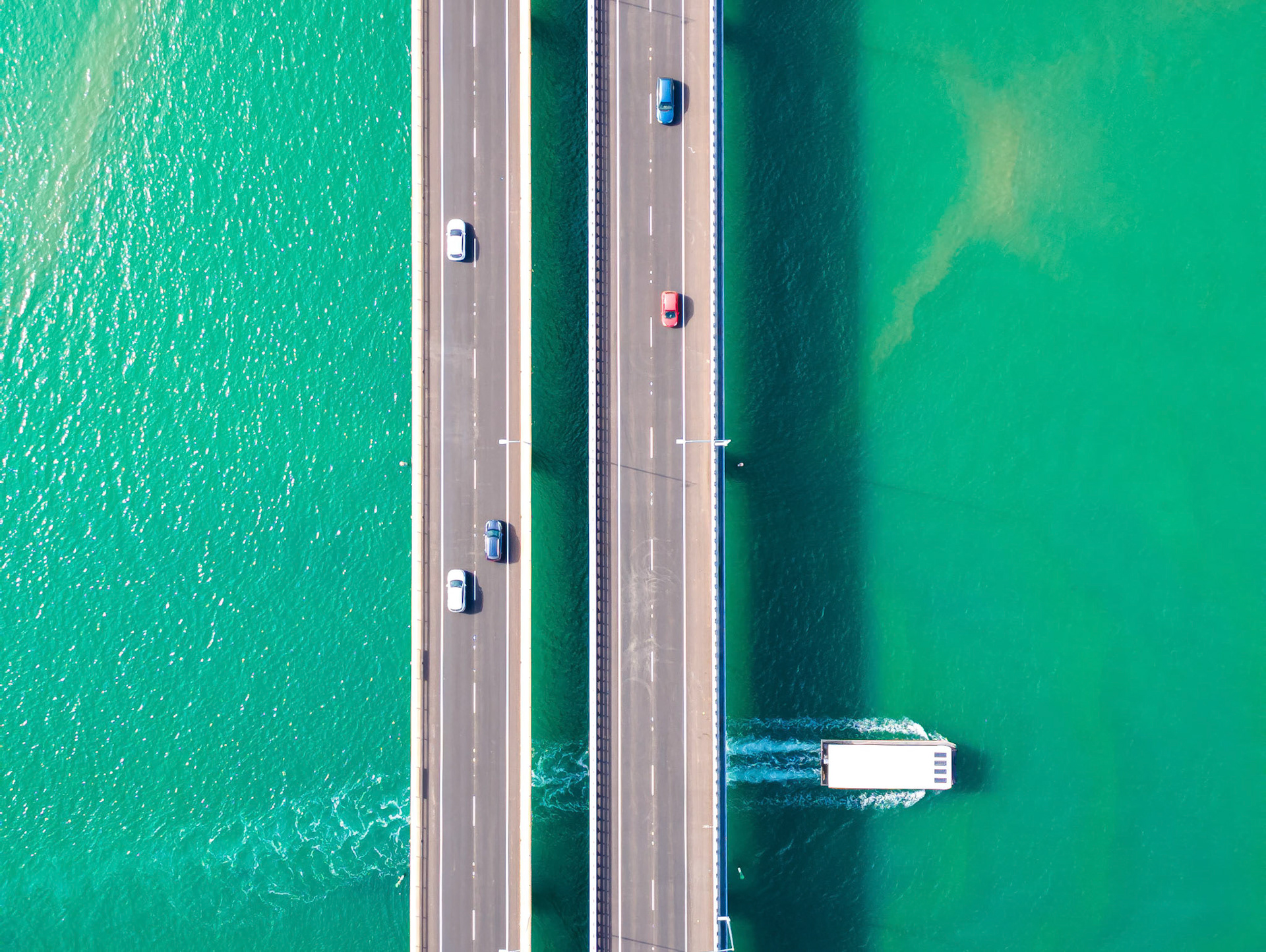 Captivating aerial shot of a modern bridge spanning over stunning turquoise waters. The image showcases multiple lanes of vehicular traffic traversing the bridge, while a boat glides beneath. The bright colors of the water contrast beautifully with the asphalt of the road, creating a visually striking composition ideal for travel, lifestyle, and transportation themes.