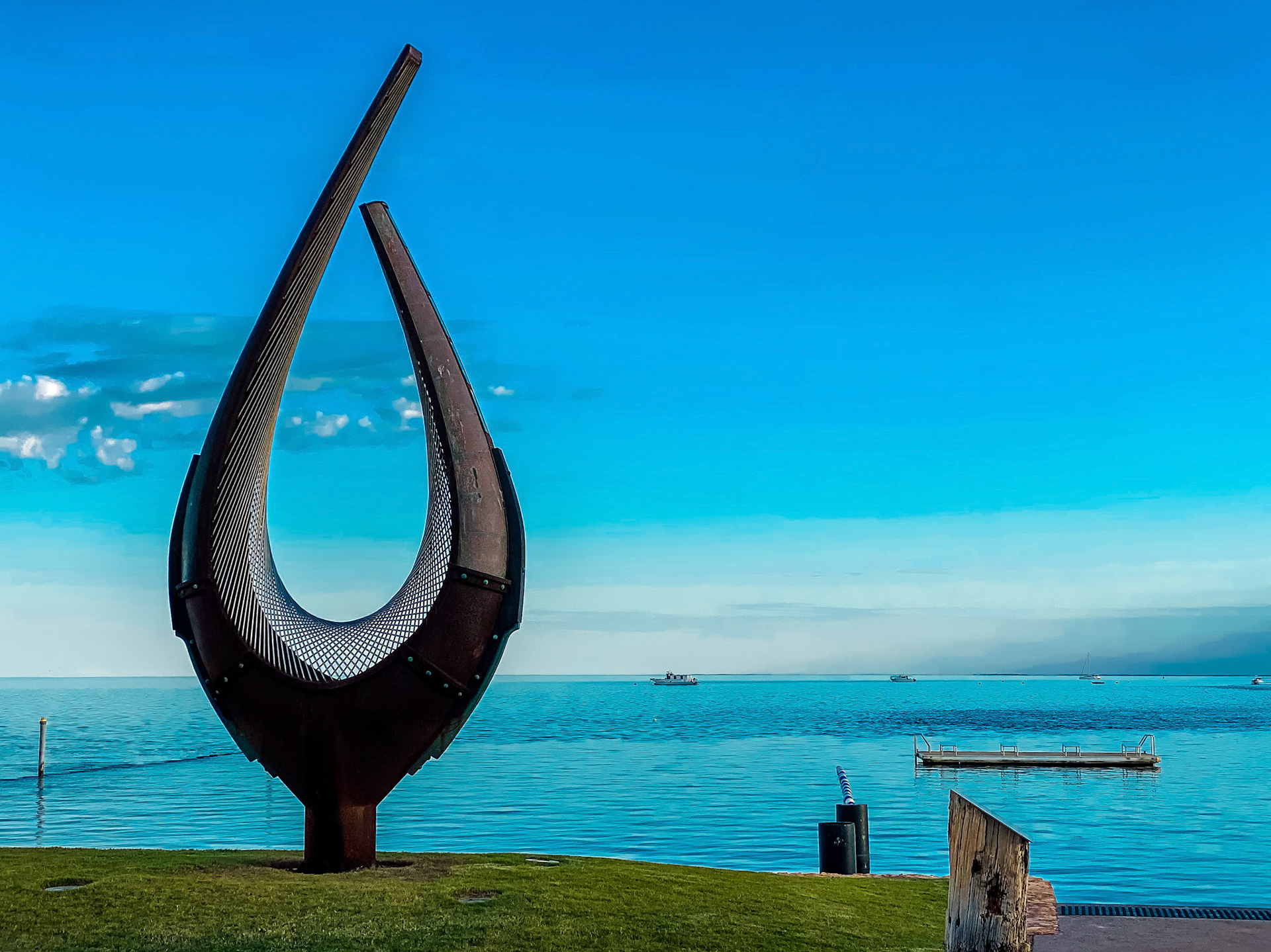 An abstract metal sculpture stands on a grassy embankment, overlooking the ocean. This modern piece enhances the coastal landscape.