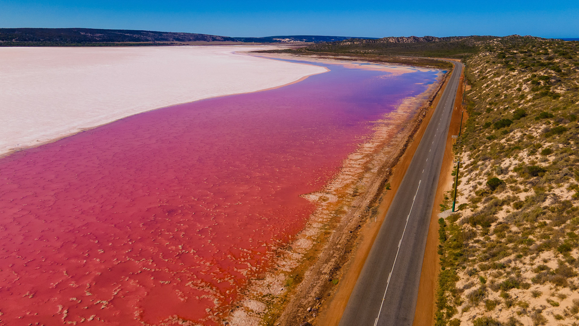 An aerial view showcases a pink salt lake alongside a solitary road, highlighting the contrast of colors in a serene natural landscape.