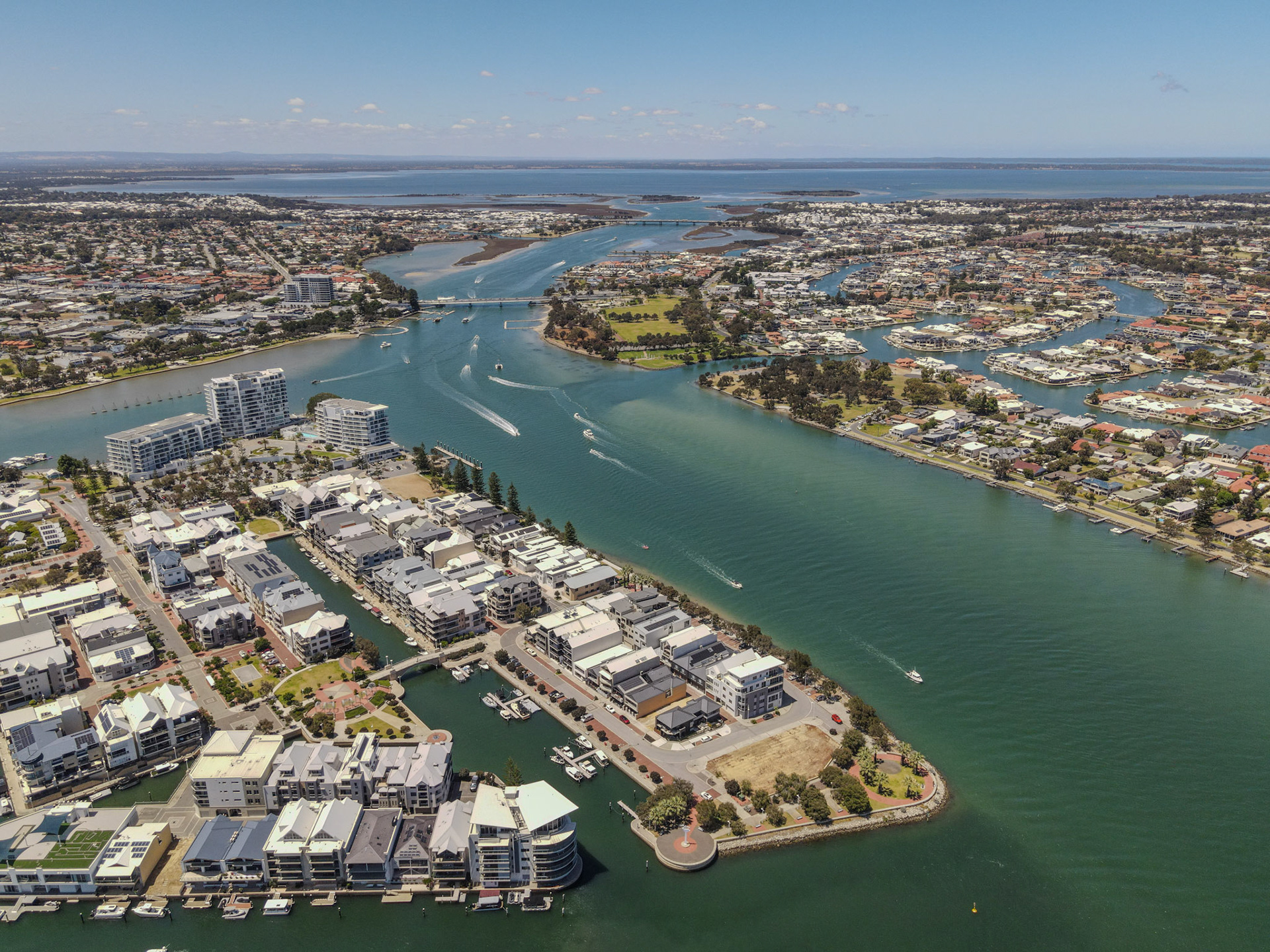 A bright, high-angle view shows a coastal city shaped by winding waterways. Boats leave white wakes along a broad green-blue channel, while a bridge spans the main junction. On one side, mid-rise apartments and marinas line the shore; on the other, curving residential canals and leafy streets spread outward. In the distance, sandbars and open water meet the horizon under a clear, sunny sky.