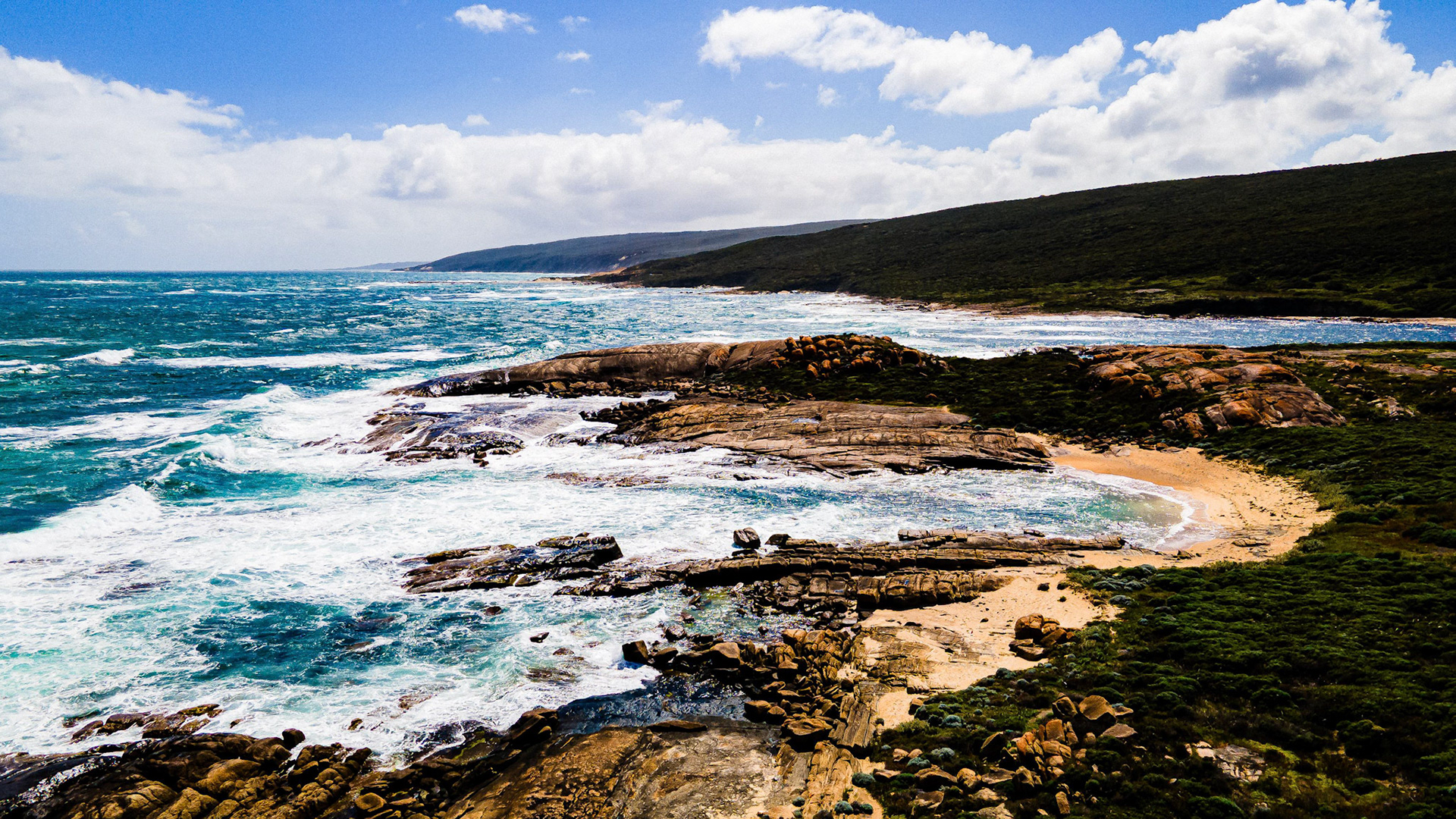 An aerial shot showcasing a stunning coastal landscape, featuring rugged cliffs and vibrant turquoise waves crashing against the shoreline. The expansive view captures the untouched beauty of nature with clear skies and rolling hills in the background, providing a picturesque scene of serenity and natural wonder.