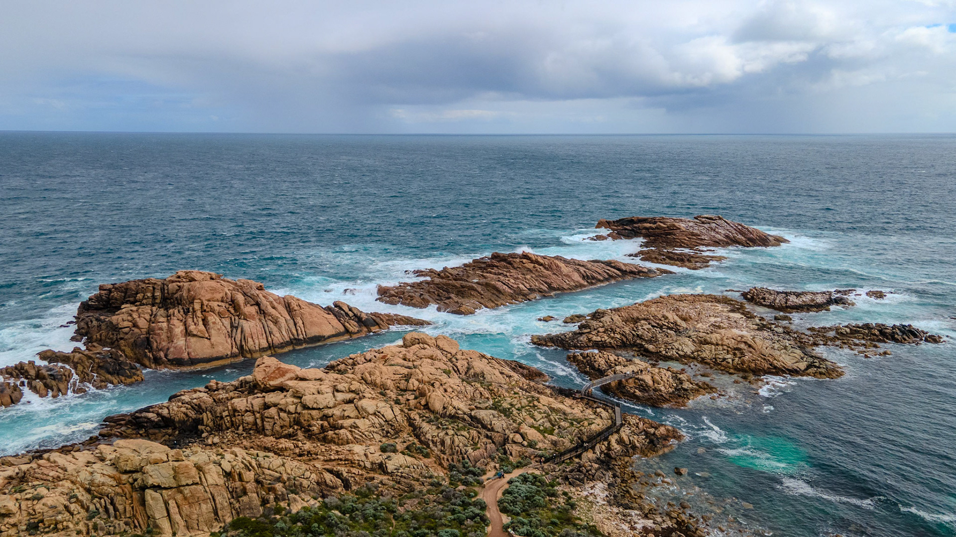 A breathtaking aerial view of a rugged coastal landscape featuring dramatic rocky outcrops extending into the vast ocean. The waves crash against the rocks, creating a dynamic contrast between the rich earth tones of the rocks and the vibrant blue hues of the sea. The cloudy sky adds depth and mood to the scene, enhancing the natural beauty of this coastal environment.