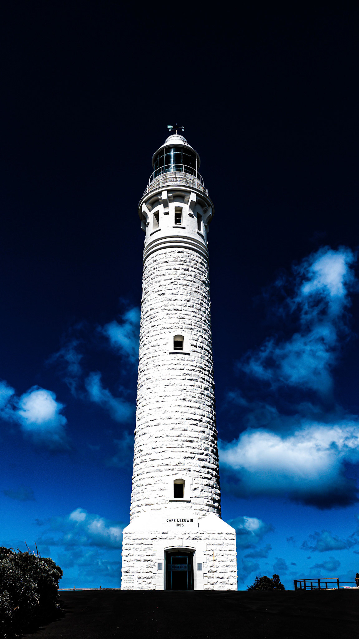 A striking image of a historic lighthouse standing tall against a vivid deep blue sky with scattered clouds. The white stone exterior contrasts beautifully with the intense sky, highlighting the architectural details of the lighthouse, built in 1895. Located at Cape Leeuwin, this iconic maritime structure serves as a beacon of navigation and a symbol of resilience along the coastline.