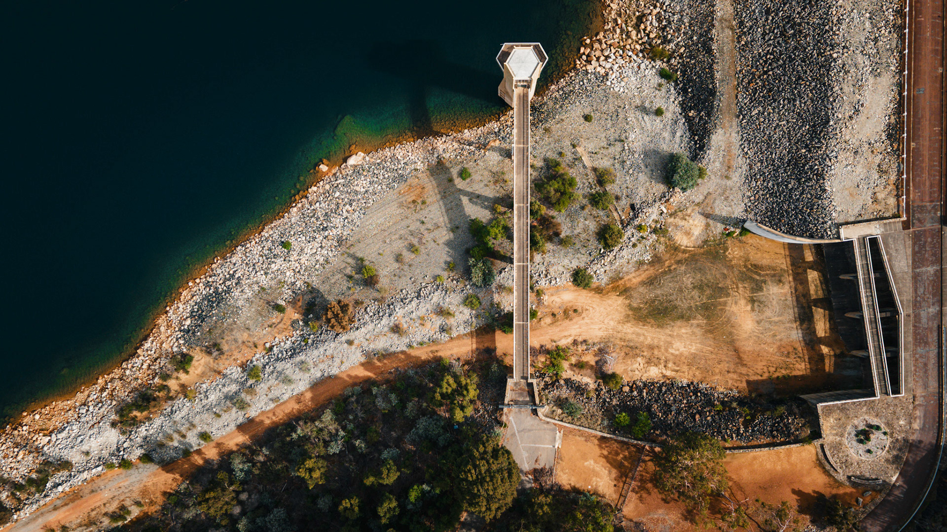 The scene shows a water reservoir with a dam. The land around the water has rocks and some plants. A tower structure is visible. Bright daylight highlights the area.