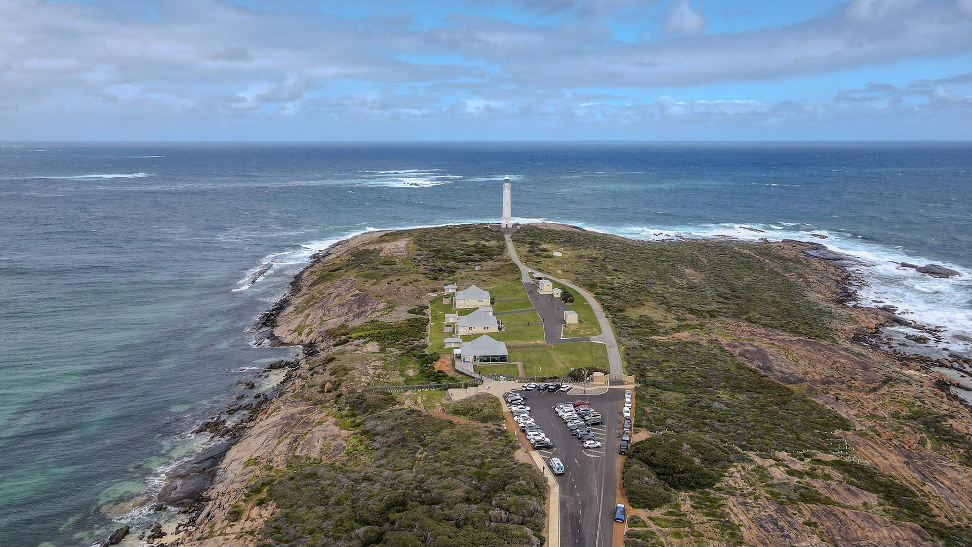 A stunning aerial view of a picturesque lighthouse situated on a rocky coastline overlooking the expansive ocean. The scene captures the natural beauty of the sea and sky, highlighted by the lighthouse's white structure. Surrounding buildings and a parking area are visible, bordered by lush greenery and rugged cliffs.