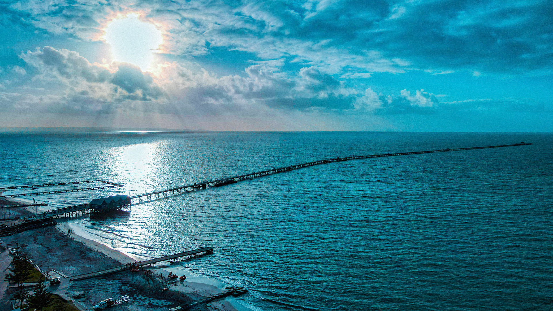 High-angle aerial view of a long pier extending into a vast ocean under a dramatic, cloud-filled sky. The sun peeks through the clouds, casting radiant light reflections on the water. The shoreline is dotted with small buildings and a few people, adding a sense of scale and tranquility to the scenic coastal landscape. The image captures the serene beauty and expansive horizon of the coastal environment, perfect for travel and nature themes.