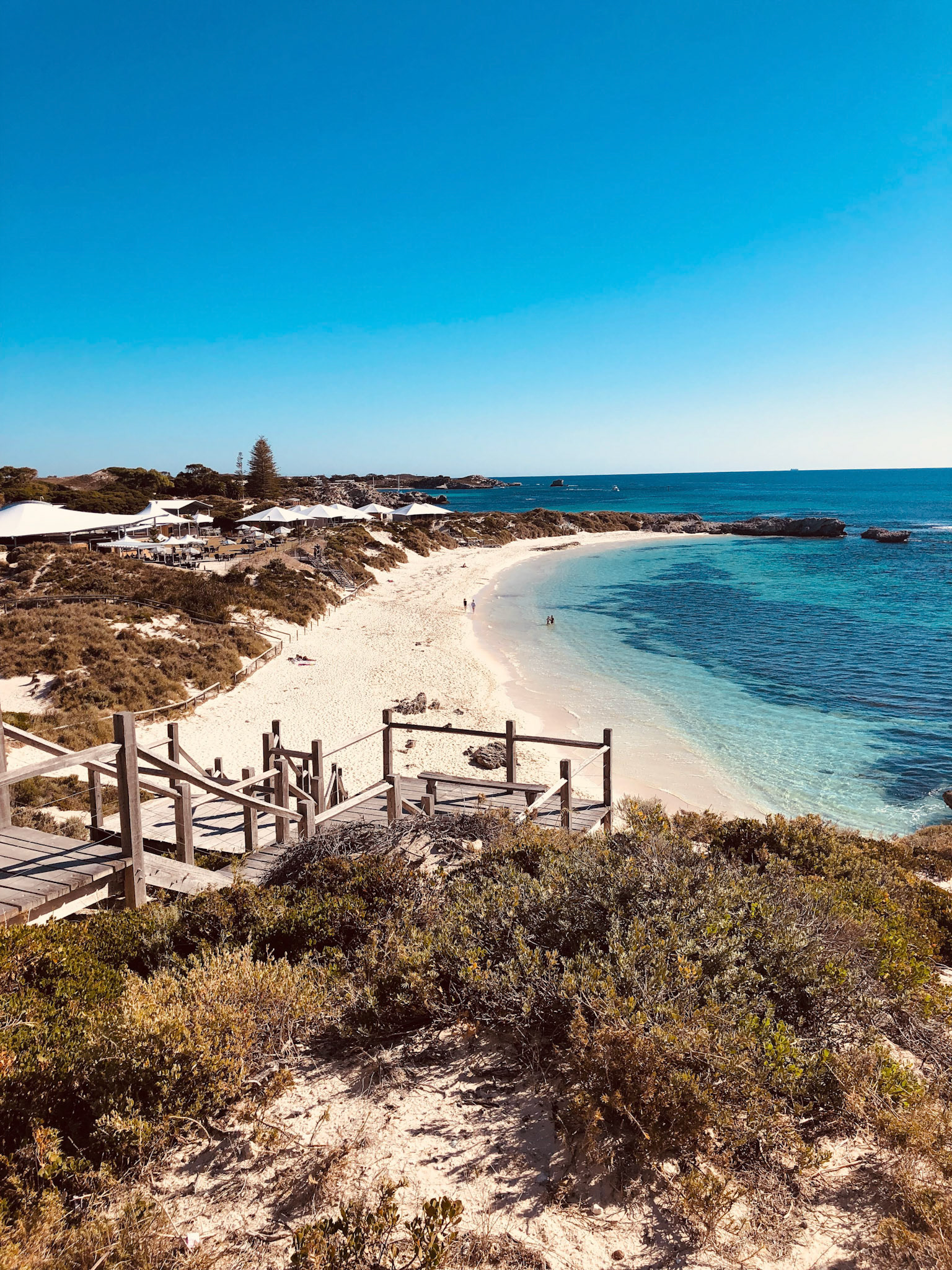 A scenic beach view features turquoise water, a wooden boardwalk, and people enjoying the sunny day. Nature and beauty abound.