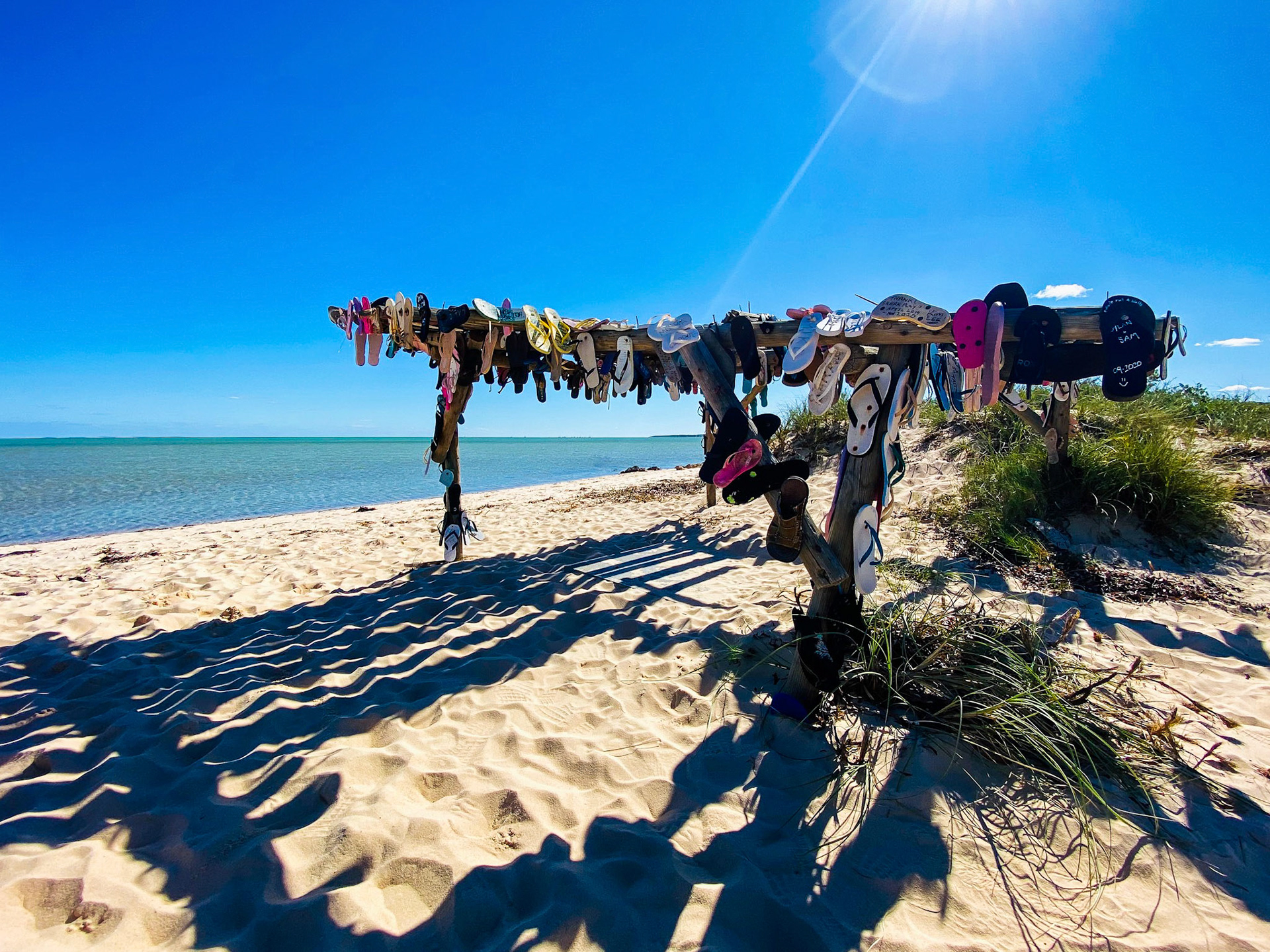 A rustic wooden structure on a sandy beach displays flip flops and hats drying under the sun, evoking summer memories and relaxation.