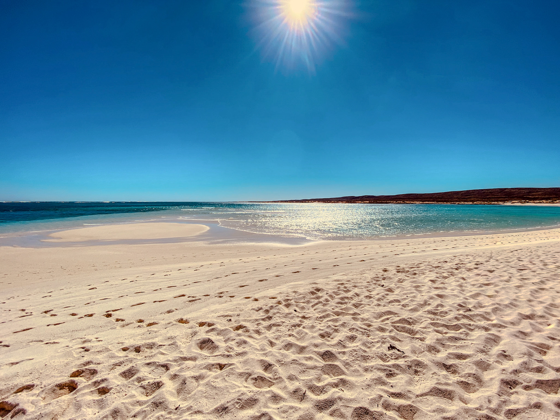 A tranquil beach features pristine white sand and a calm turquoise ocean under a bright sky. Footprints hint at recent visitors.