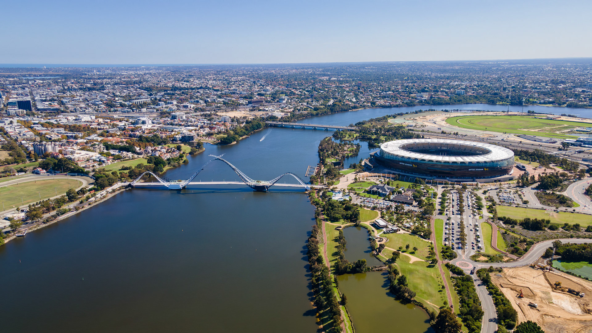 Majestic skyline reveals a bustling city with a grand stadium beside a winding river, surrounded by serene parks. Bright sunlight illuminates the entire scene, creating a lively atmosphere.