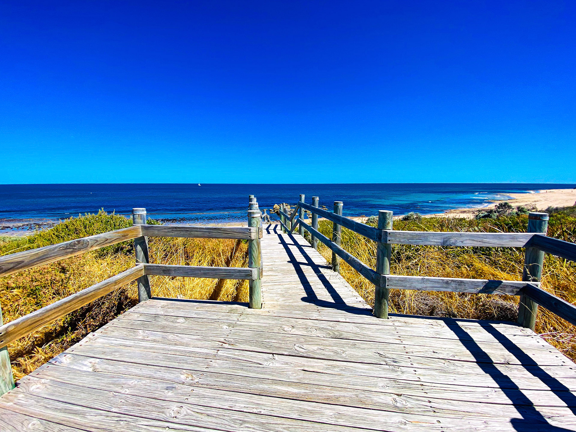 A weathered boardwalk leads to a tranquil beach and deep blue ocean under a clear sky. This scene embodies summer serenity.