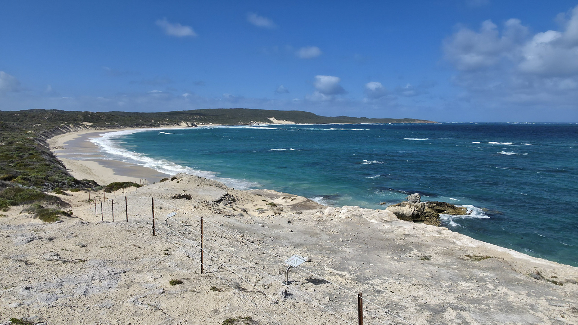 A picturesque view of a pristine ocean shoreline featuring crystal-clear blue waters against a backdrop of rugged, rocky terrain and lush greenery. The beach stretches into the distance under a vibrant blue sky dotted with fluffy clouds, creating a tranquil and inviting landscape perfect for travel and tourism representations.
