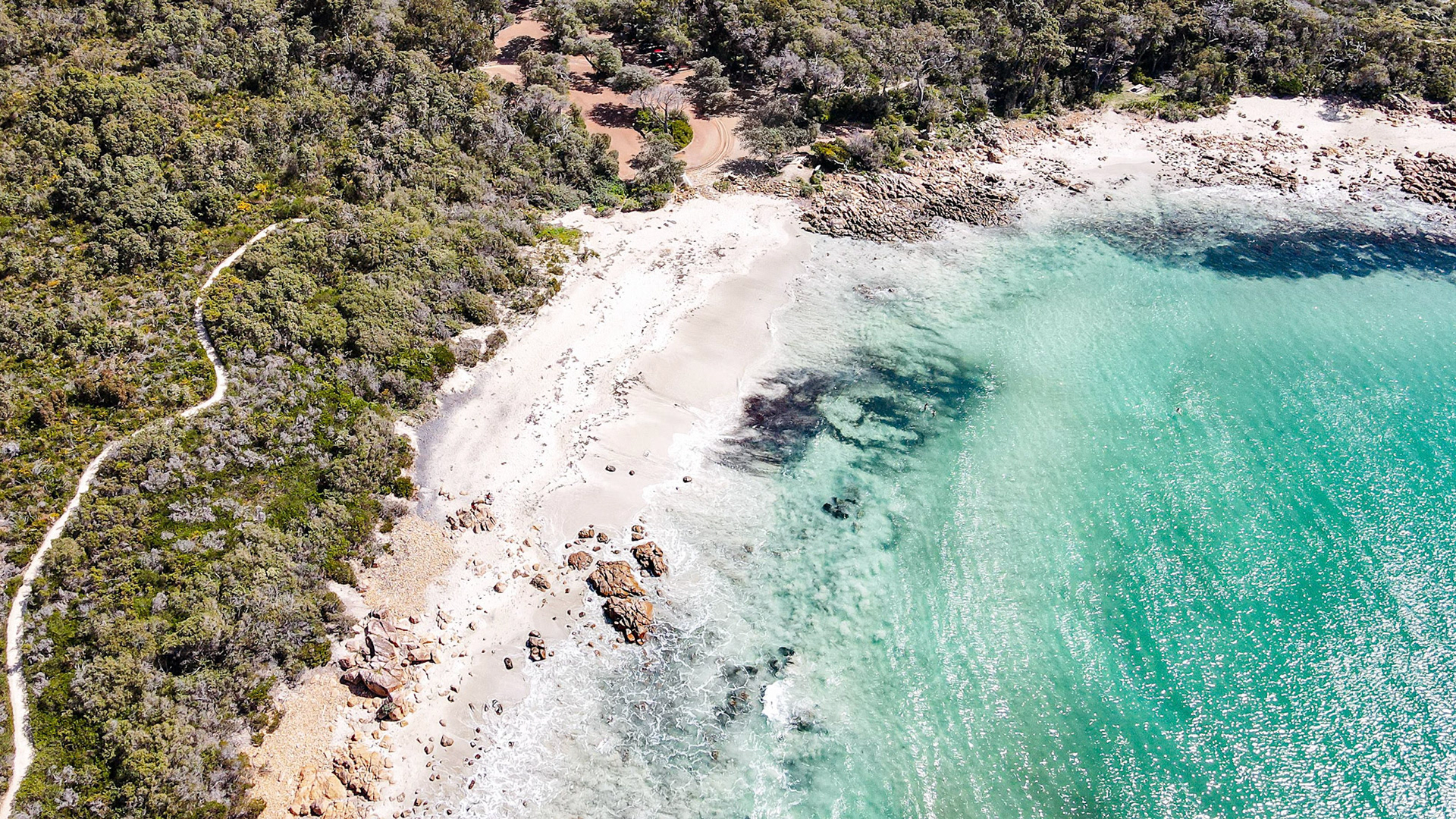 A stunning aerial shot captures the natural beauty of a secluded beach with fine white sand and rocky outcrops nestled beside lush green foliage. The vibrant turquoise waters of the ocean gently lap against the shore, creating a tranquil and picturesque scene ideal for nature lovers and travel enthusiasts. The winding path through the greenery adds a sense of adventure and exploration to the idyllic landscape.