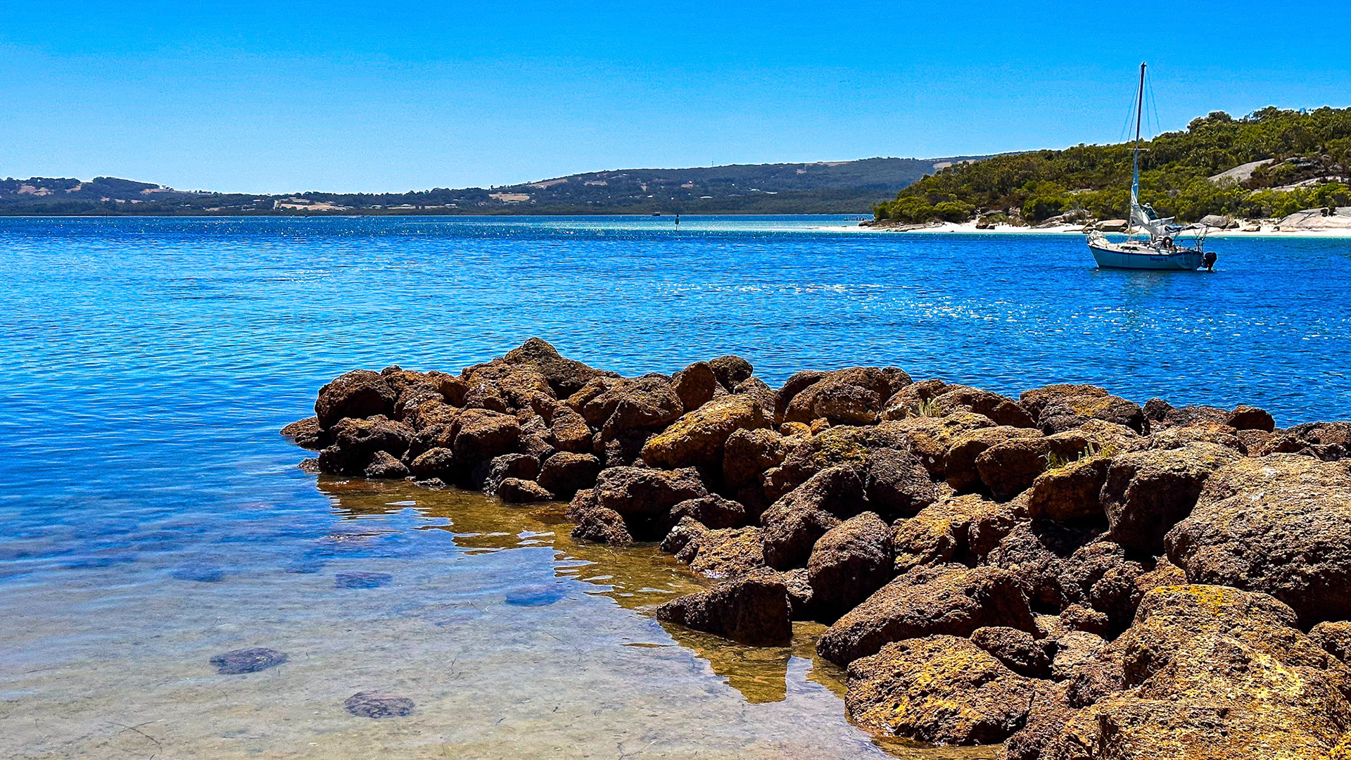 A sailboat floats on the vibrant ocean, surrounded by rocks and trees under a clear sky, creating a serene coastal scene.
