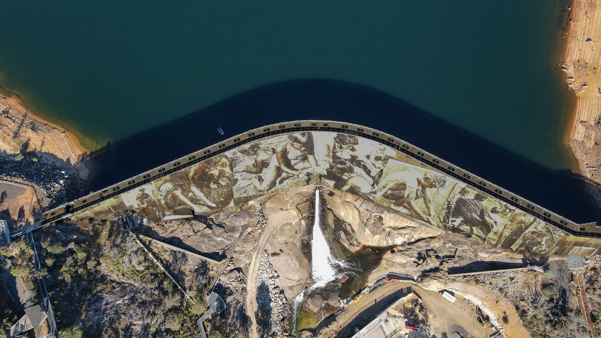 A dramatic top-down view of a curved concrete dam holding back deep teal water. The dam wall is covered by a giant sepia-toned mural depicting people at work and at rest. Below the wall, a bright white plume pours from the spillway into a rocky gorge, with access roads, buildings, and rugged terrain surrounding the structure. A small boat floats near the dam on the reservoir side, and terraced shorelines frame the scene.