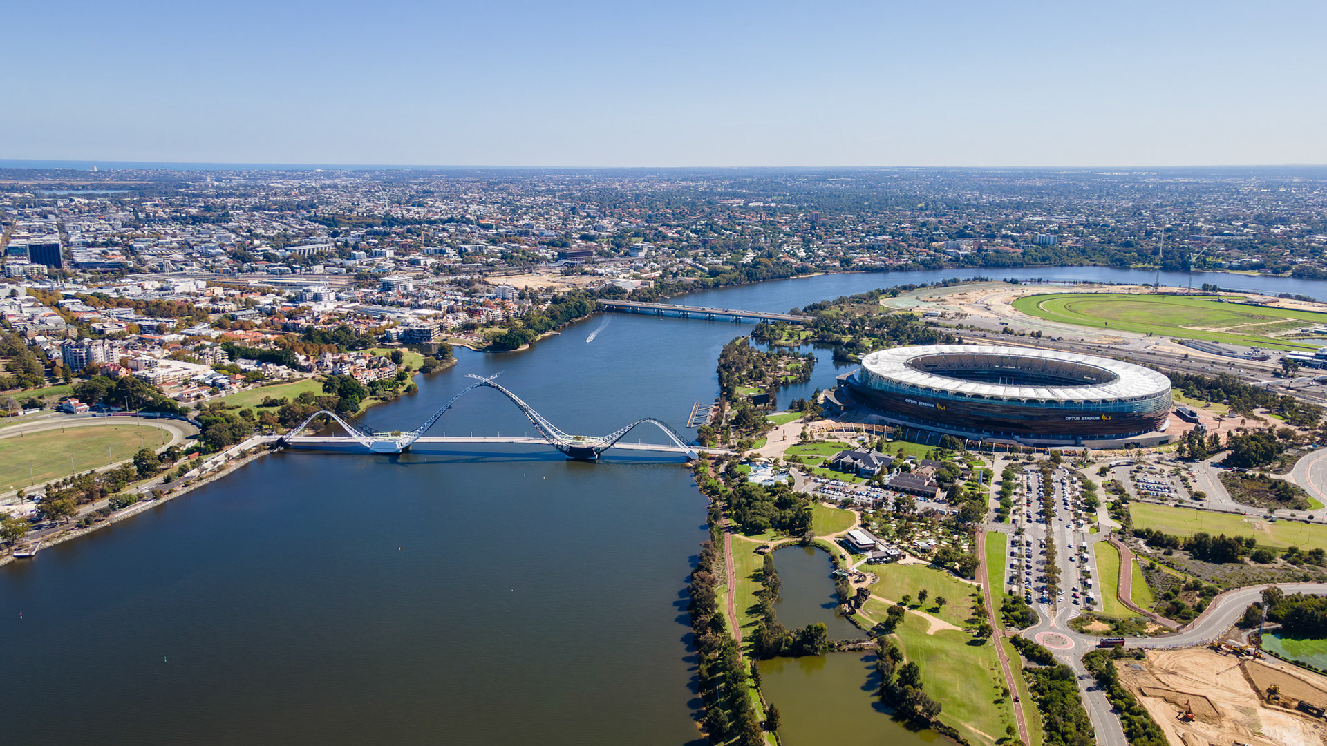A breathtaking view captures the harmony of city and nature, highlighting a modern stadium, a scenic river, and lush green parks on a sunny day. Bridges connect vibrant areas, bustling with activity.
