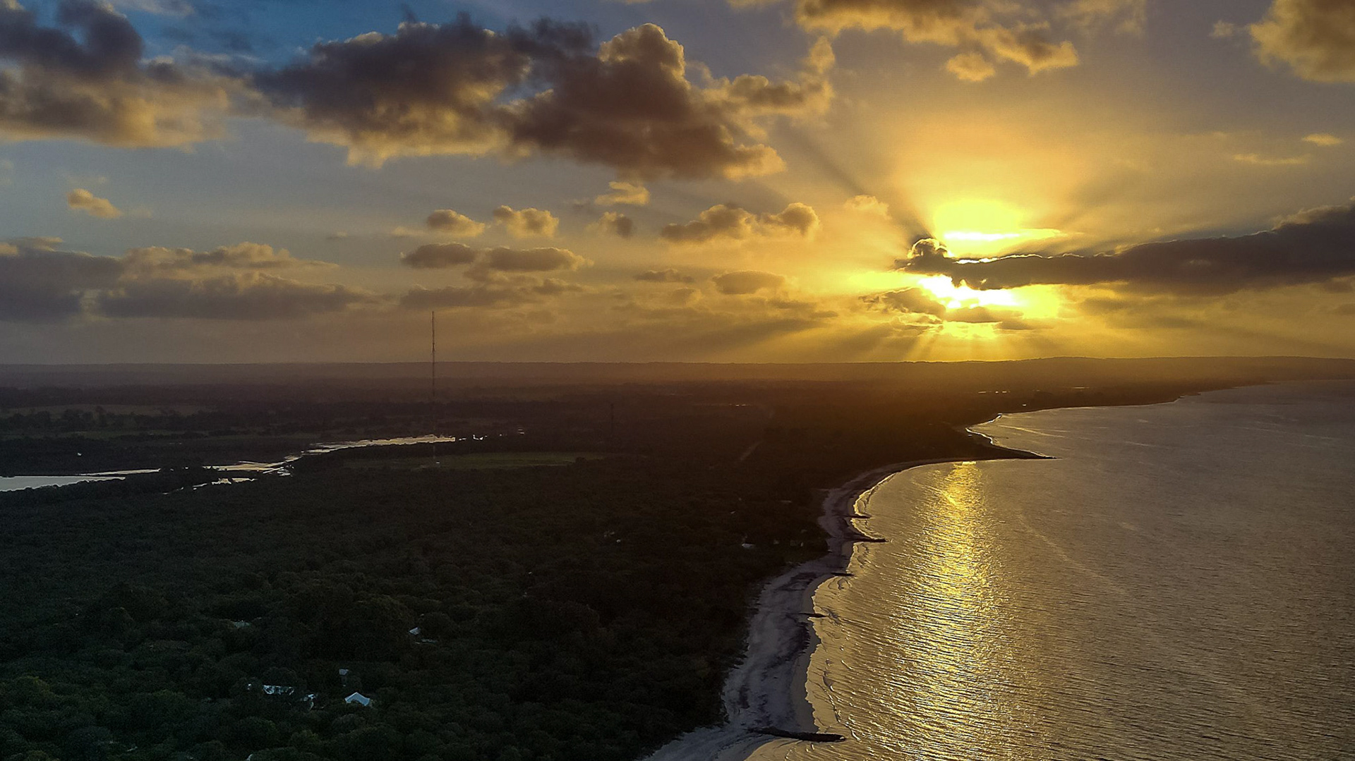 A breathtaking aerial view capturing the golden hues of a sunset over a serene coastline. The glowing sun casts radiant beams through scattered clouds, illuminating the ocean and surrounding lush greenery. This scenic image embodies the tranquil beauty of nature at dusk.
