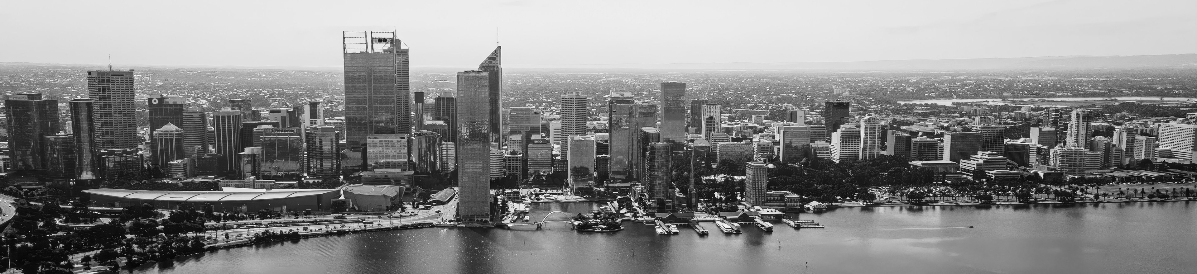 Skyscrapers rise majestically against a clear sky, reflecting sunlight on the calm river. Boats gently bob in the water, creating a serene urban scene.