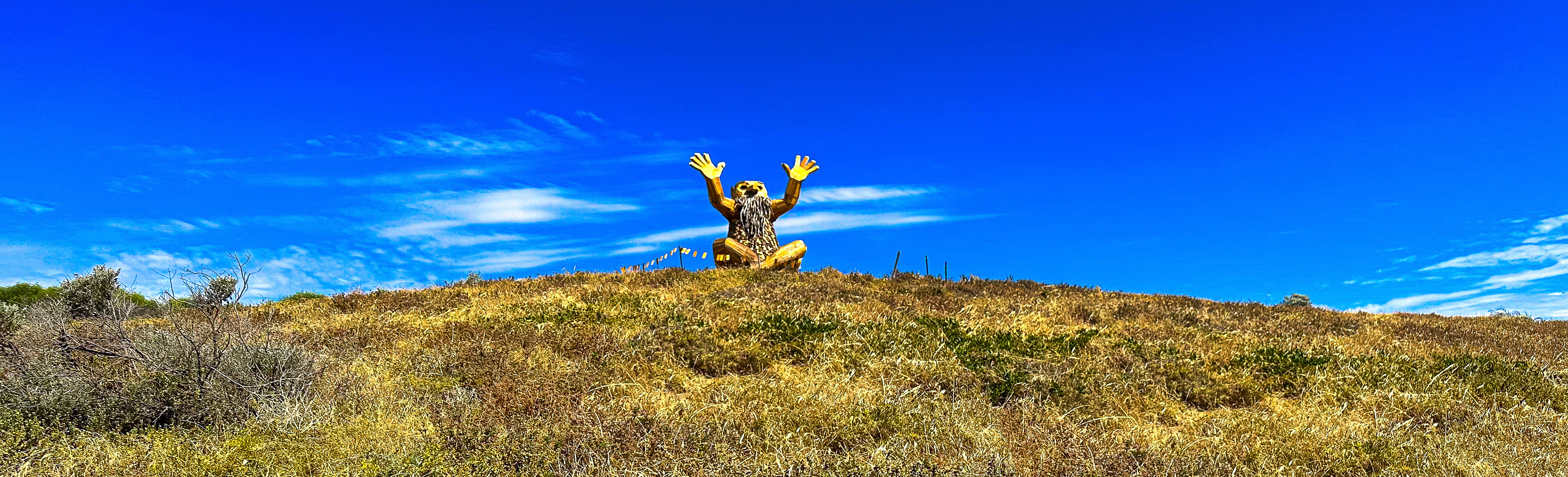 A figure sits cross-legged on a grassy hilltop, arms raised in joy against a clear blue sky, embodying freedom and connection with nature.