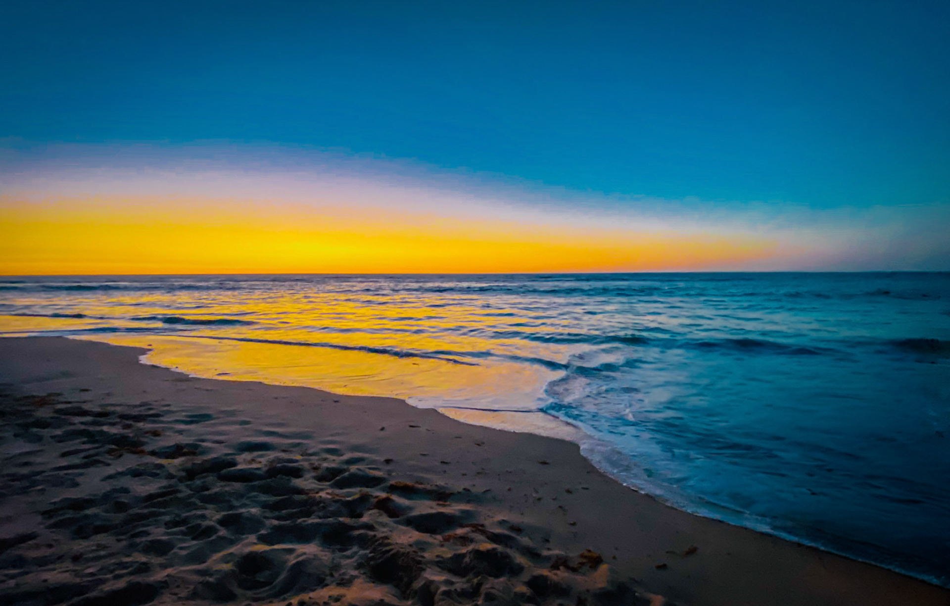 A vibrant sunset creates a serene atmosphere with warm hues over the ocean. Waves gently lap at the sandy shore, reflecting tranquility.