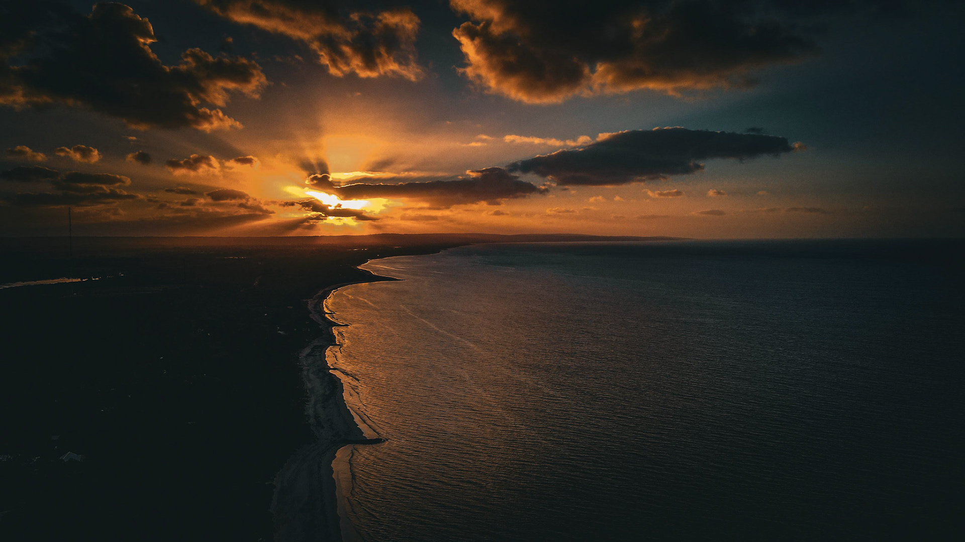 Captivating aerial view capturing a breathtaking sunset over a serene coastline. The vibrant orange and deep blue hues blend seamlessly as the sun sets on the horizon, casting dramatic shadows over the ocean waves and rugged shoreline. Wispy clouds accentuate the sky, creating an awe-inspiring vista perfect for travel and nature enthusiasts.