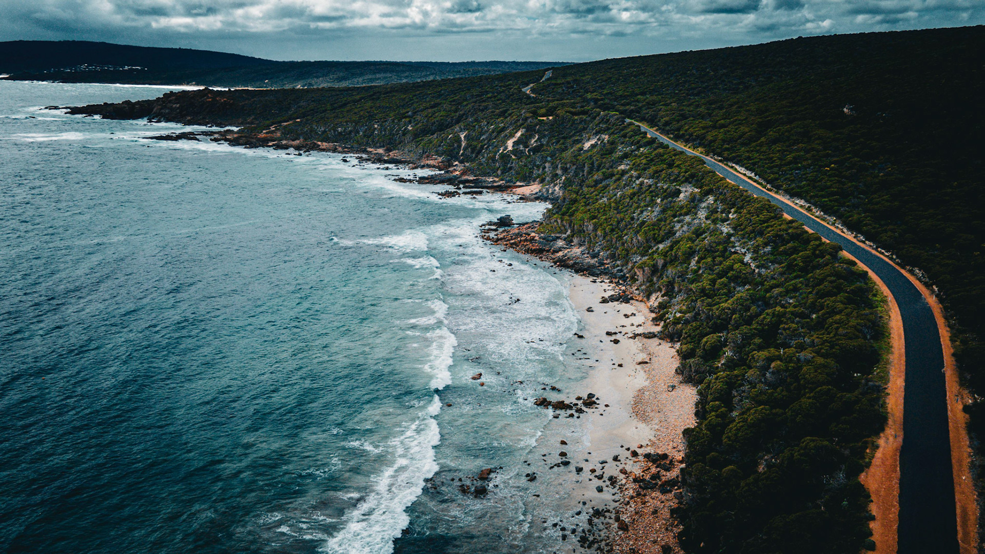 An aerial view captures the stunning contrast between a winding coastal road and a rocky beach with turquoise waters. Dense green foliage covers the adjacent cliffs, and waves gently lap against the shoreline under a cloudy sky, creating a serene and picturesque landscape perfect for travel and nature enthusiasts.