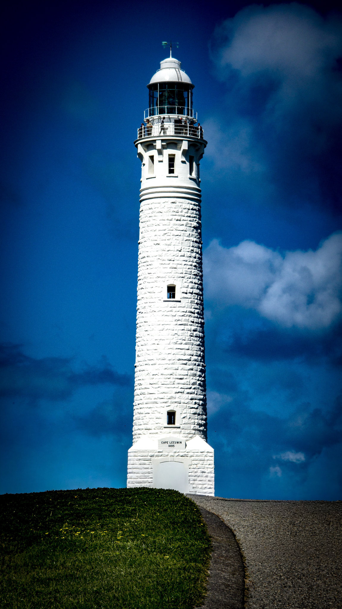 A striking lighthouse stands tall against a vibrant blue sky with scattered clouds, creating a scenic coastal view. The lighthouse's white facade contrasts beautifully with the sky, emphasizing its architectural elegance and historical significance. The foreground features a gently sloping grassy hill, adding a natural element to the composition.