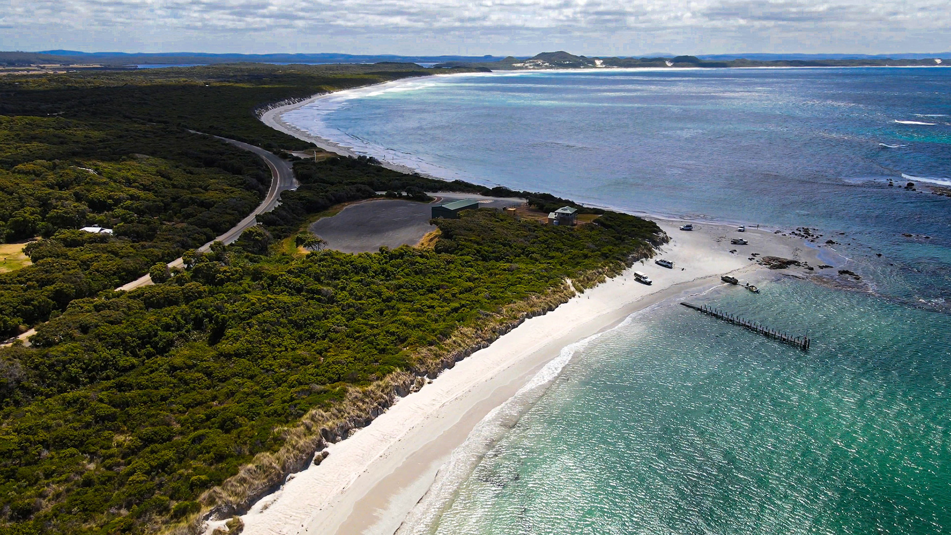 An aerial perspective showcases a tranquil coastal landscape featuring a white sand beach, lush vegetation, and the vast blue ocean.