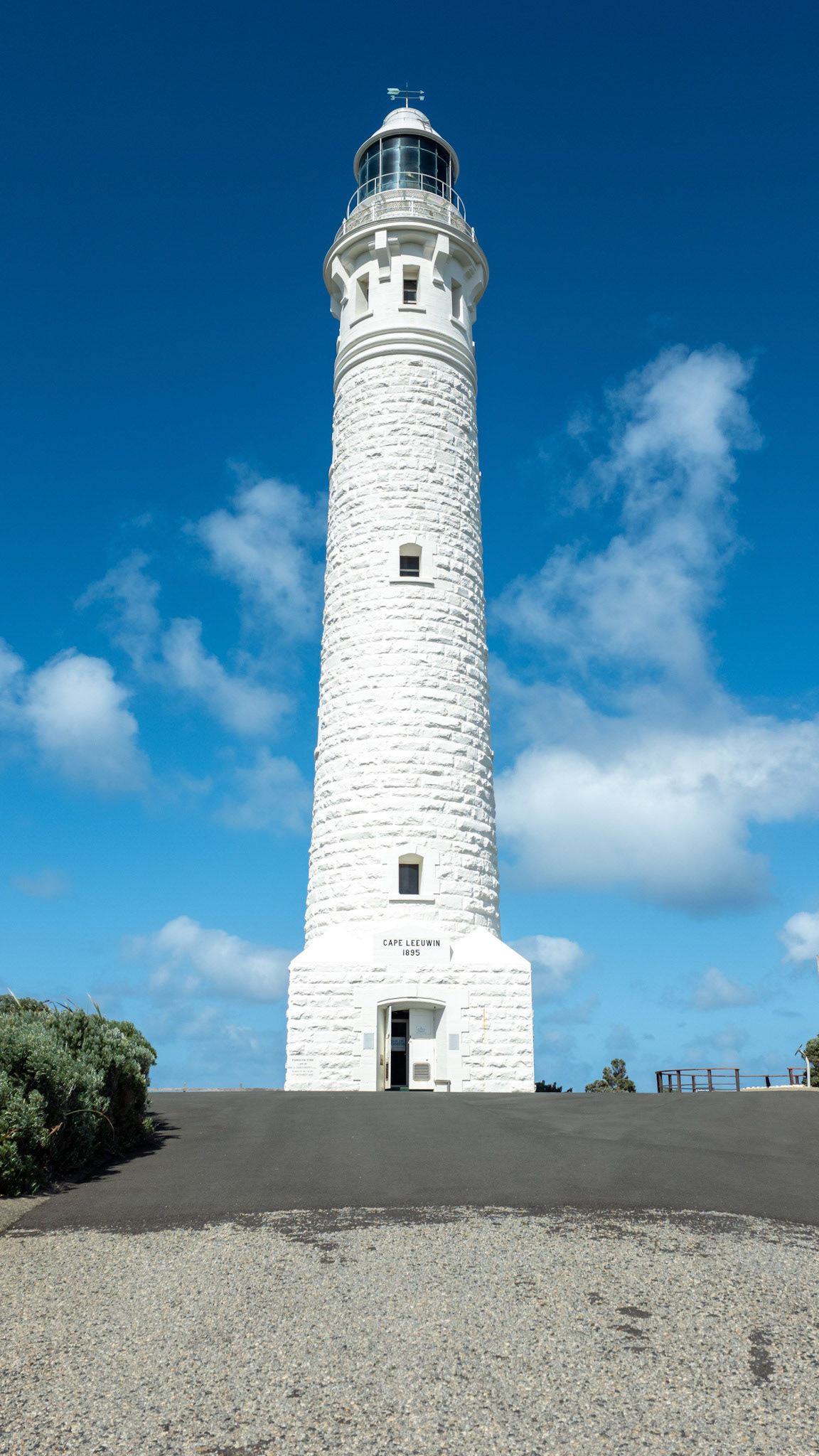 A stunning stock image showcasing a towering white lighthouse set against a vivid blue sky, offering an awe-inspiring view. The lighthouse, positioned centrally in the frame, exhibits its iconic structure and historical significance, making it a perfect representation of architectural beauty by the sea. The low angle shot emphasizes the lighthouse's height and grandeur, while the fluffy clouds add depth and interest to the scene. Ideal for themes of navigation, coastal architecture, and travel.