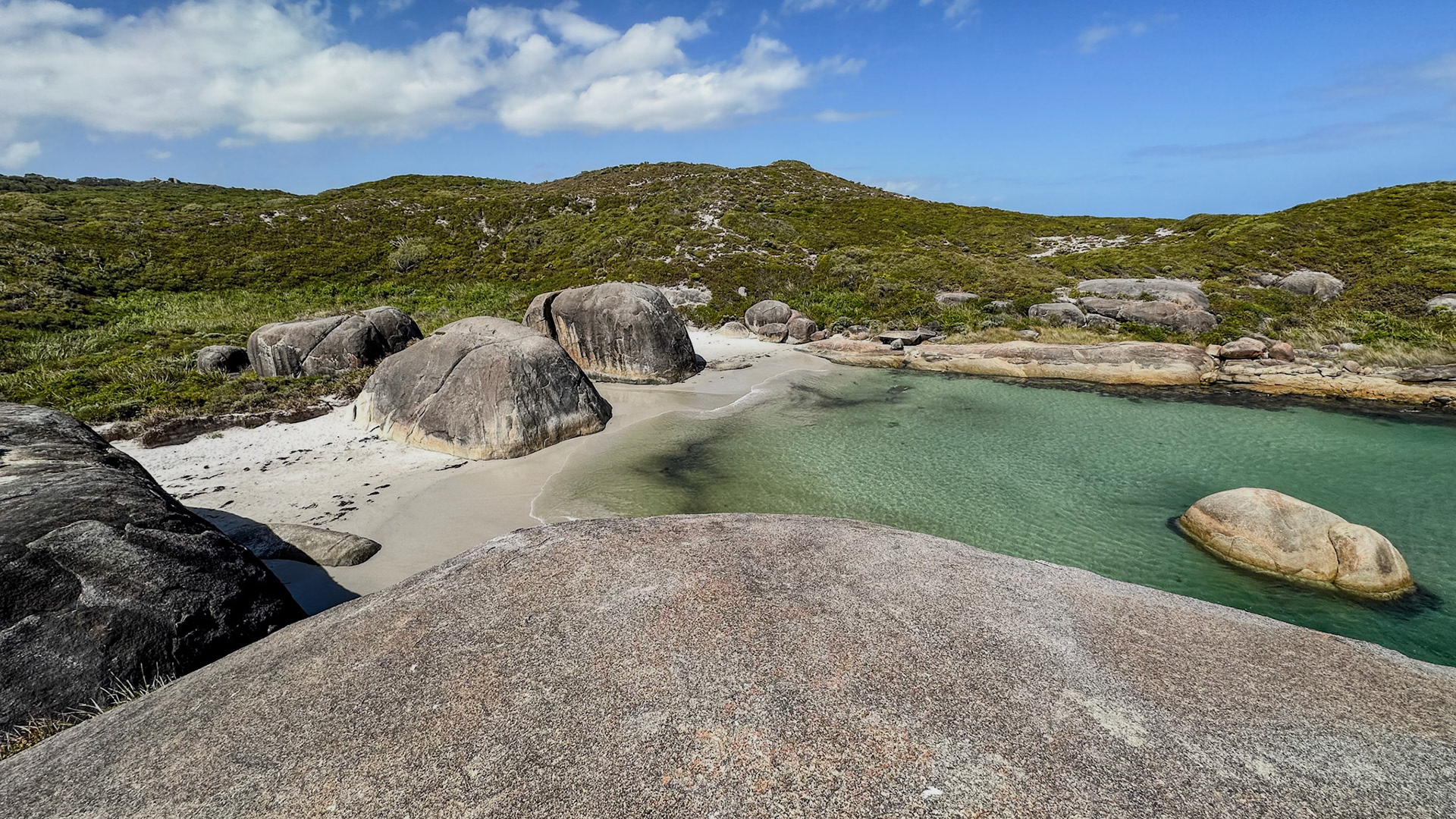 A serene coastal landscape features weathered boulders on a tranquil beach, with turquoise water lapping against the shore.
