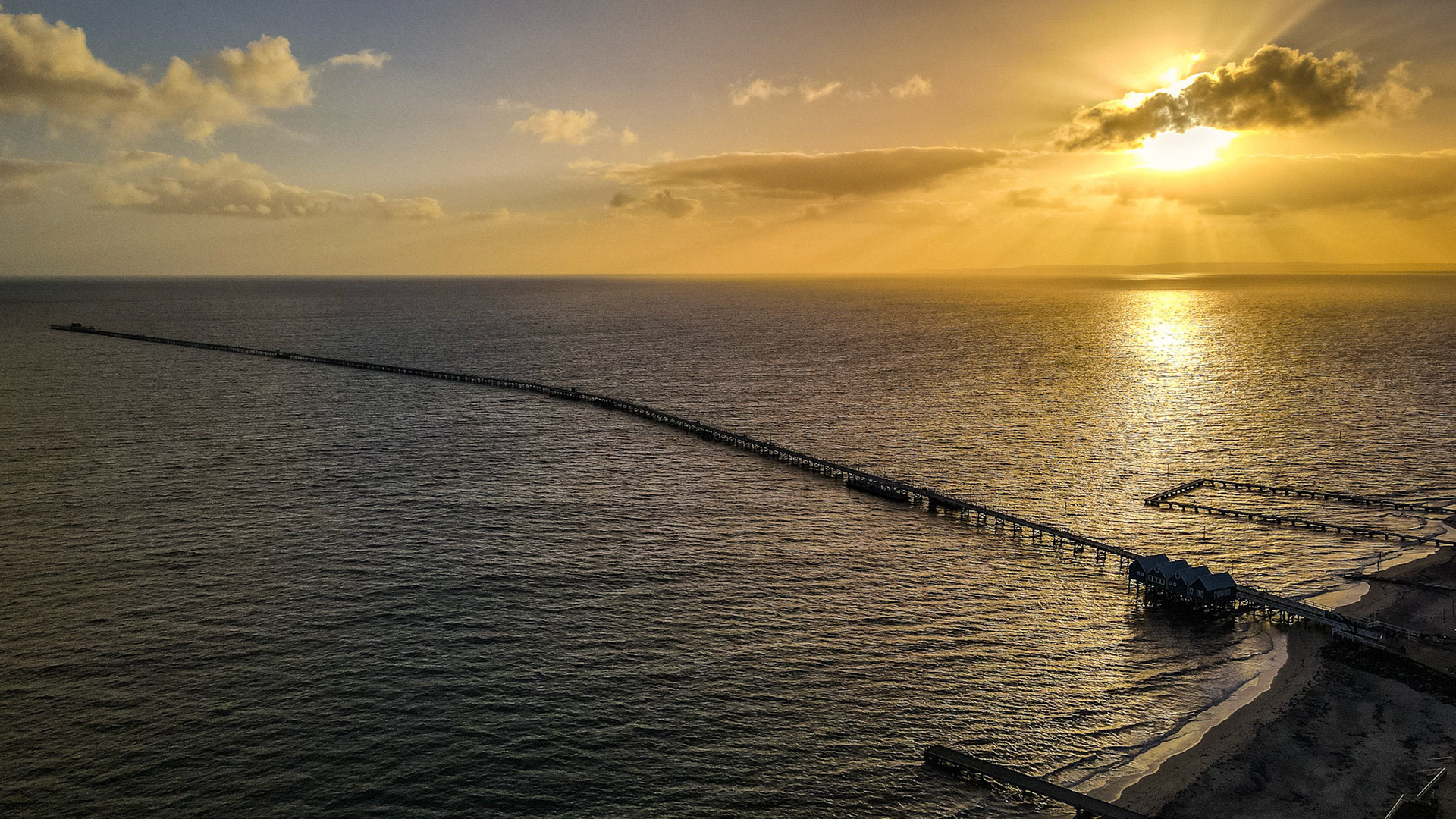A breathtaking aerial view captures a long pier extending into the ocean, bathed in the golden hues of a vibrant sunset. The sun's rays pierce through scattered clouds, casting a warm glow across the rippling water, while the serene sky creates a tranquil atmosphere. This scenic seaside landscape evokes a sense of peace and wonder, ideal for travel and nature visuals.