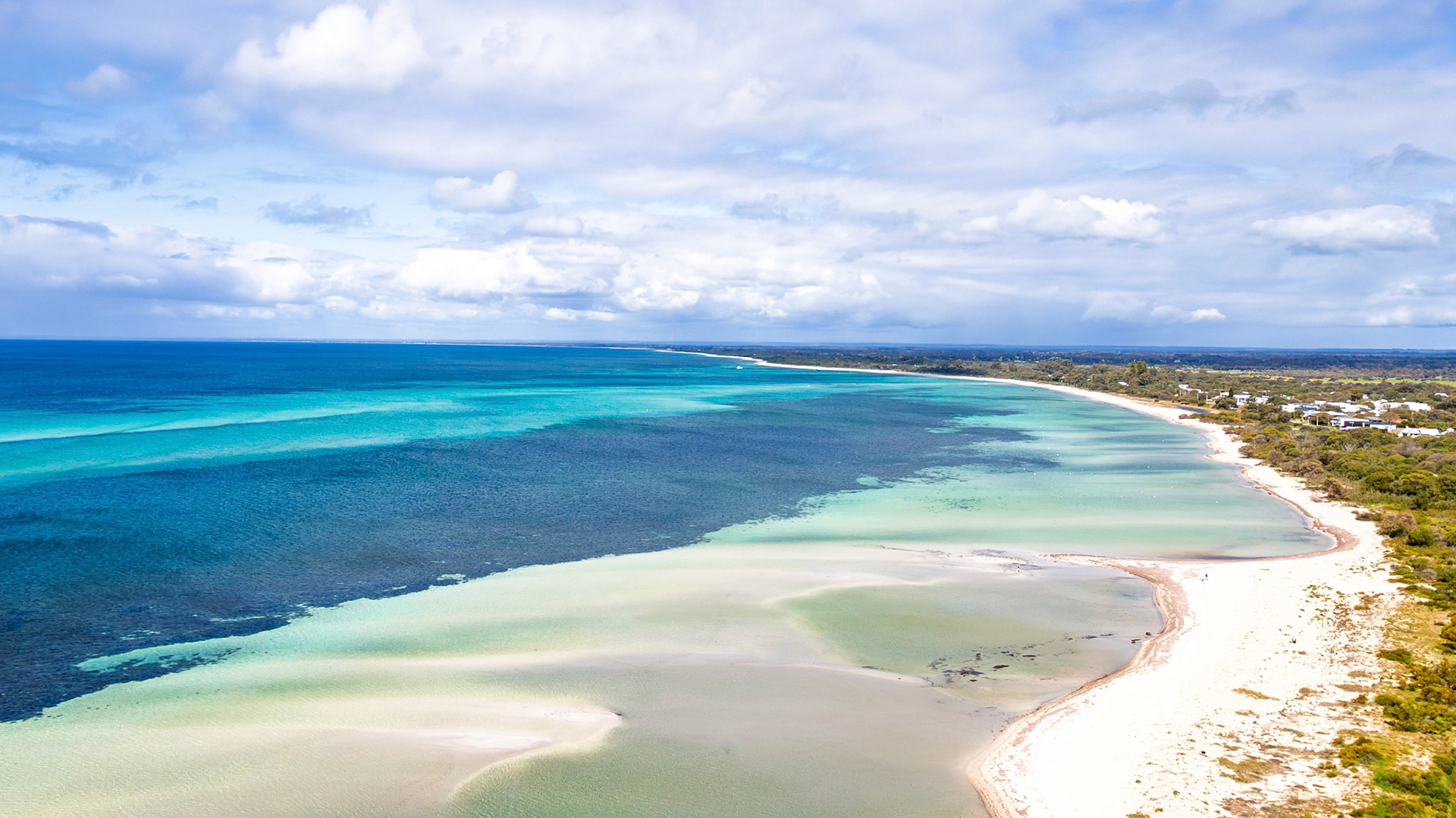 Stunning aerial view of a pristine coastline showcasing turquoise waters and sandy shores. The expansive ocean seamlessly meets the lush greenery lining the coast under a partly cloudy sky, creating a serene and picturesque landscape.