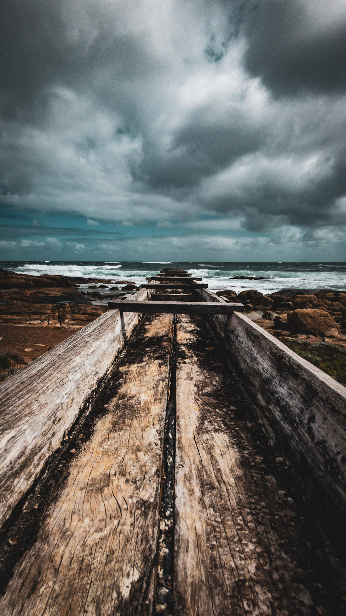 Dramatic stock image of a weathered wooden pathway extending towards the ocean, under a sky filled with moody, overcast clouds. The rugged textures of the wood contrast with the churning ocean waves in the background, capturing a sense of adventure and rugged beauty. This image evokes feelings of exploration and the raw power of nature.