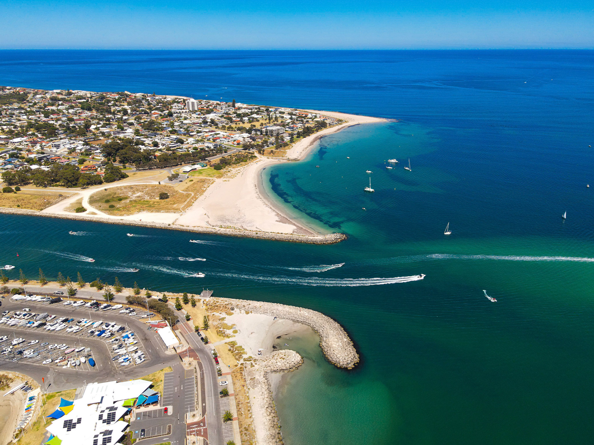 A wide aerial view captures a sweeping coastline with a curving sandy beach, a sheltered channel bordered by rock breakwaters, and a busy marina. Speedboats and jet skis carve bright white wakes while sailboats glide in calm, turquoise water. A residential neighborhood spreads along the shore beneath a clear blue sky.