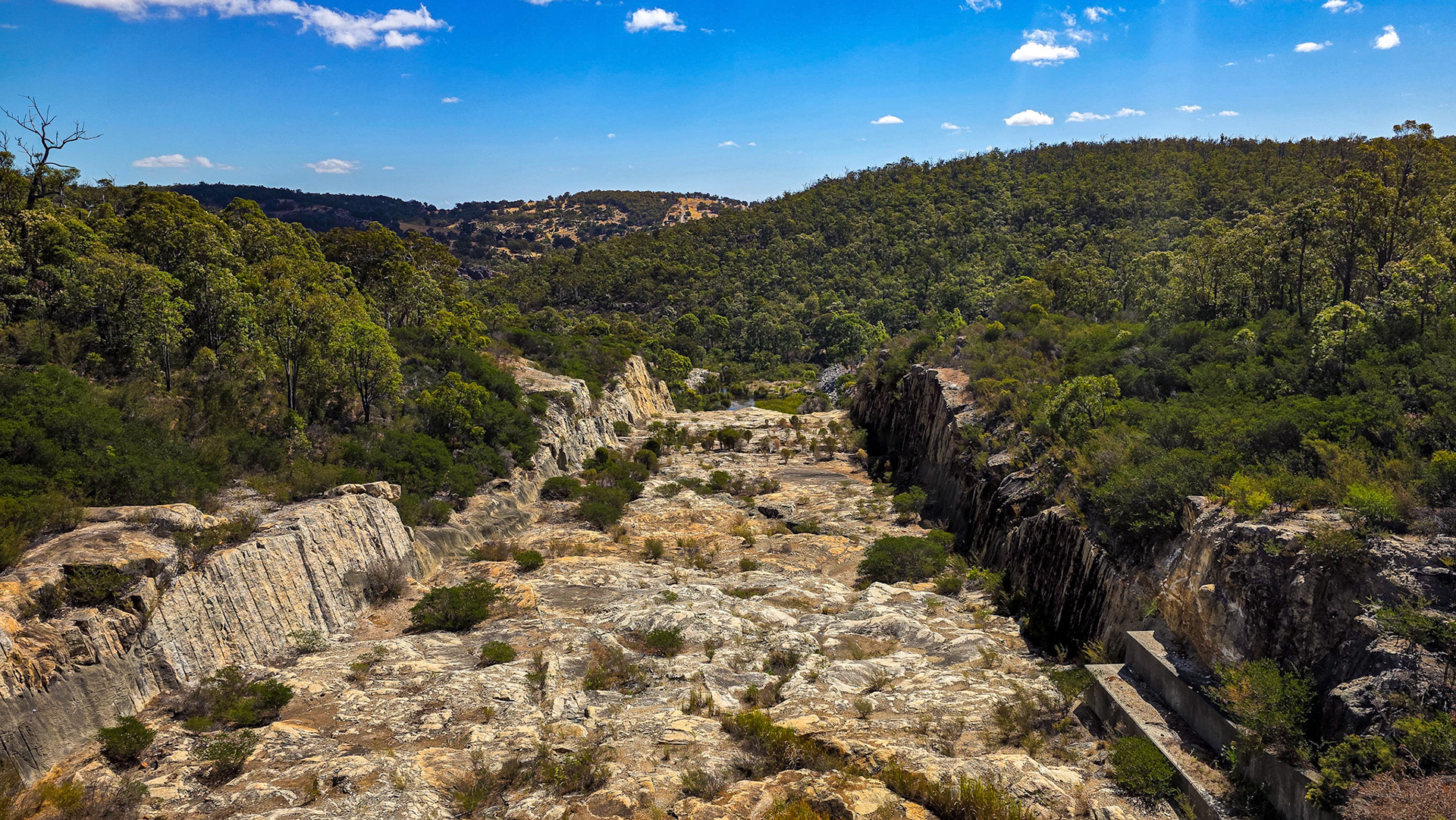 A rugged, sun-bleached gorge slices through dense green bushland, bordered by steep rock walls and scattered shrubs on the dry riverbed. Forested hills of tall trees rise on both sides under a vivid blue sky dotted with small clouds. A small concrete channel is visible at the lower right, hinting at past water flow or management.