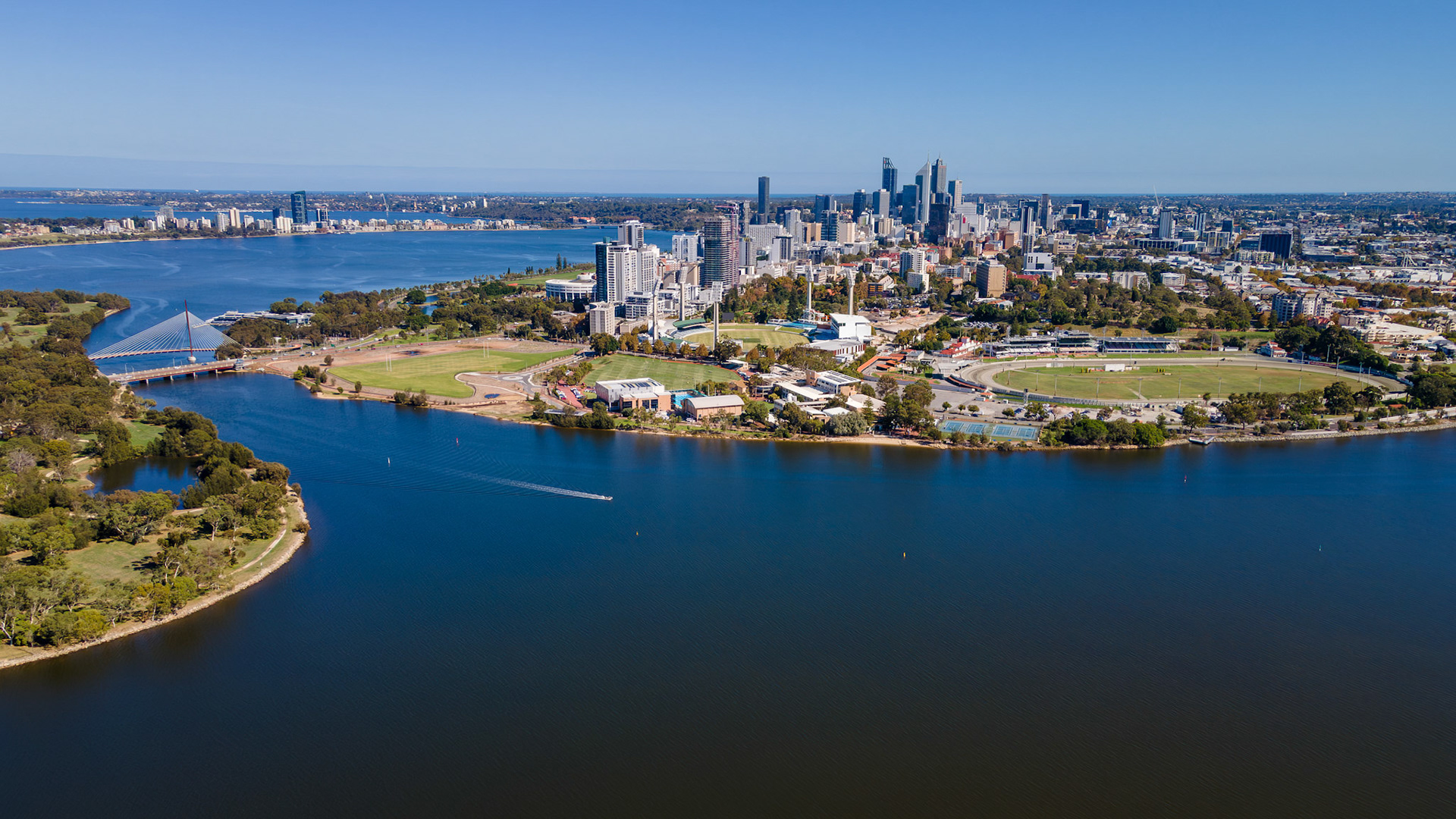 Bright sunlight bathes the stunning skyline of the city with tall buildings, while the tranquil Swan River reflects the clear blue sky, surrounded by green parks and inviting nature.
