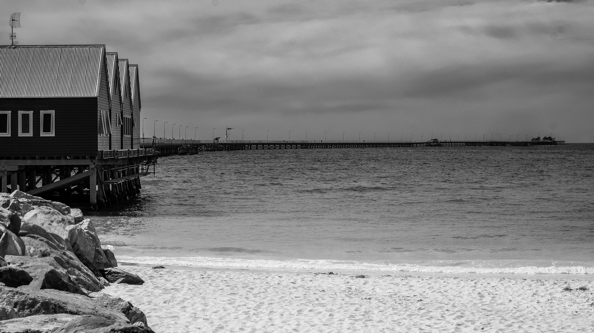 A serene black and white scene of historic wooden buildings on stilts over calm water, connected by a long pier.