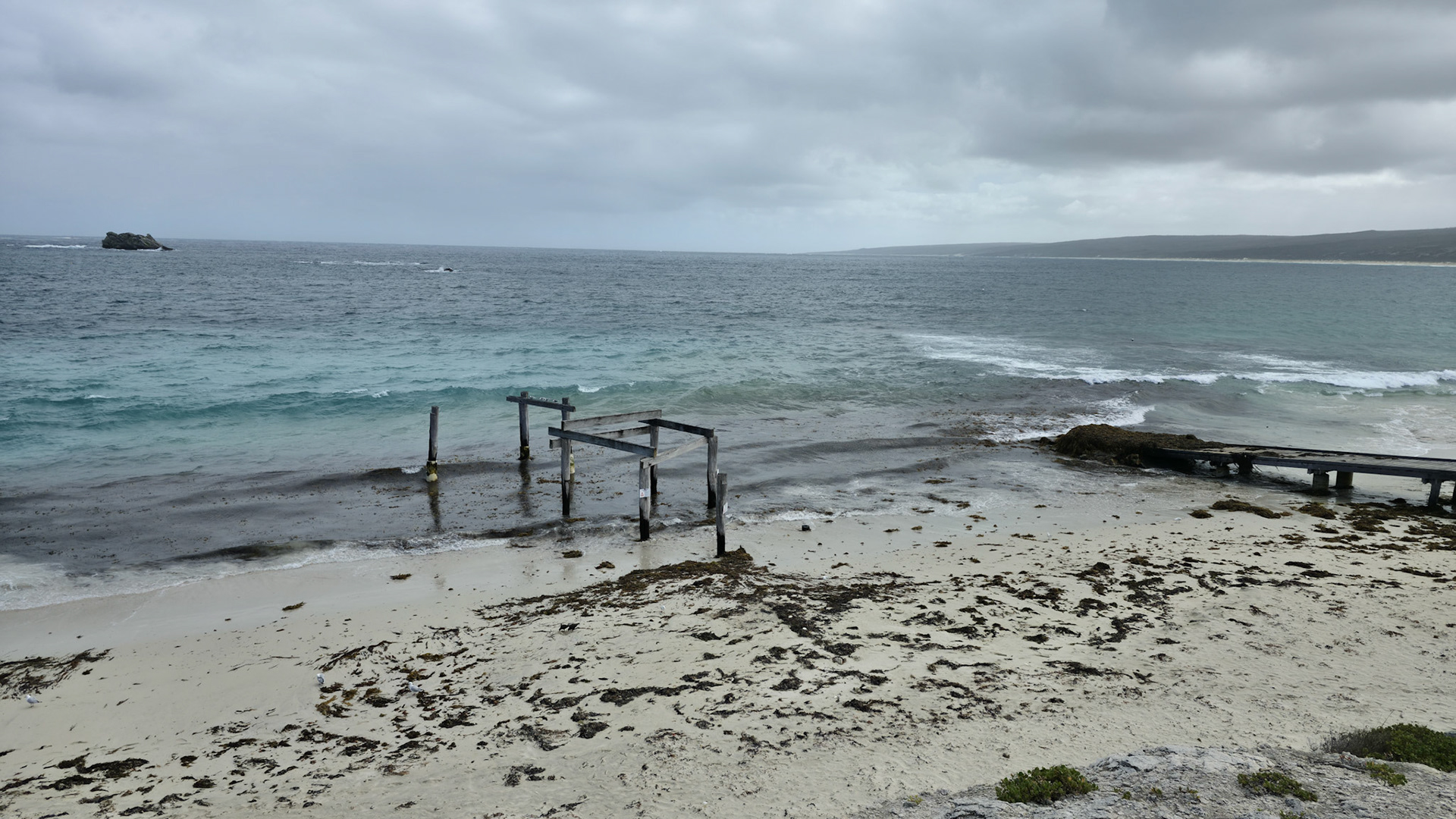 A serene coastal scene depicting an abandoned wooden pier on a sandy beach, with the ocean waves gently crashing against the shore. The cloudy sky adds a dramatic touch to the tranquil setting, highlighting the contrast between the weathered wood and the vibrant sea. Ideal for themes of isolation, nature, and the passage of time.