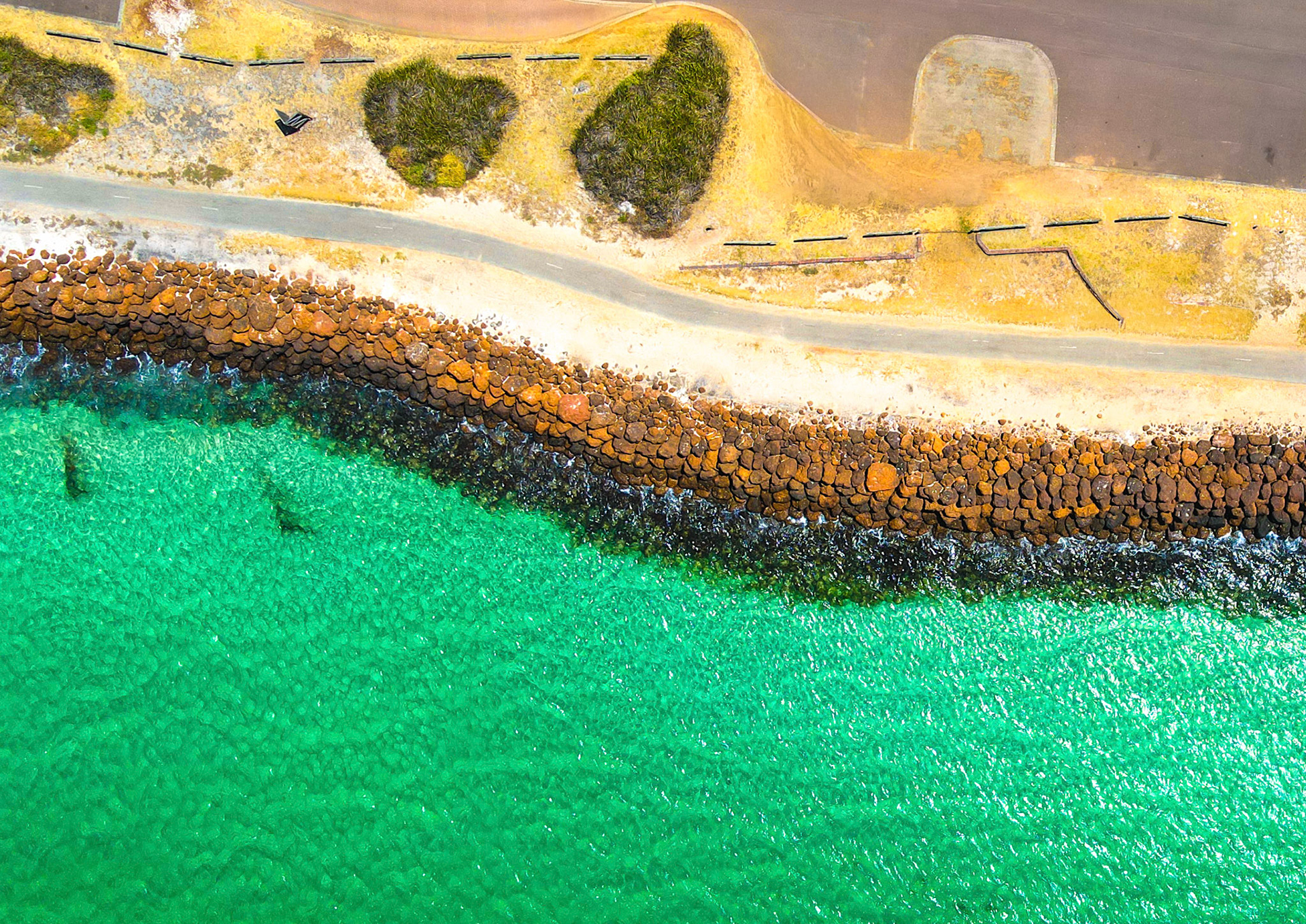 Explore a stunning aerial view of a vibrant coastline, featuring a breakwater, clear water, and a scenic path bordered by grass.