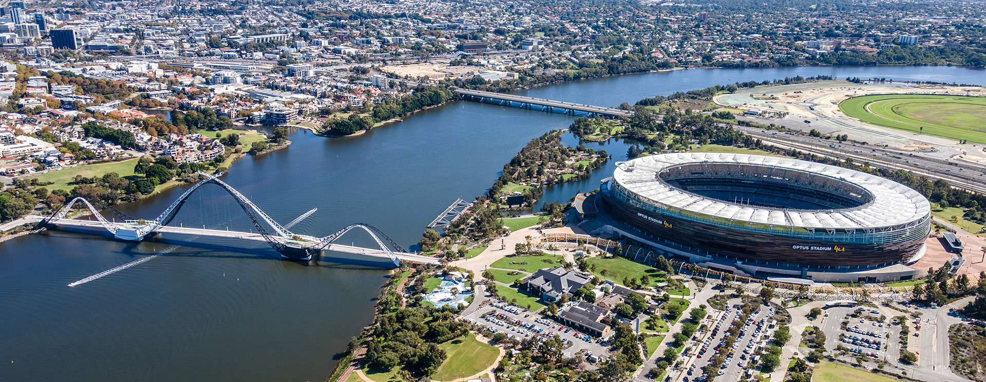 A breathtaking view showcases a sleek stadium nestled by a clear river against the backdrop of a lively city. Nearby, a graceful bridge arches over the water, connecting urban life and nature.