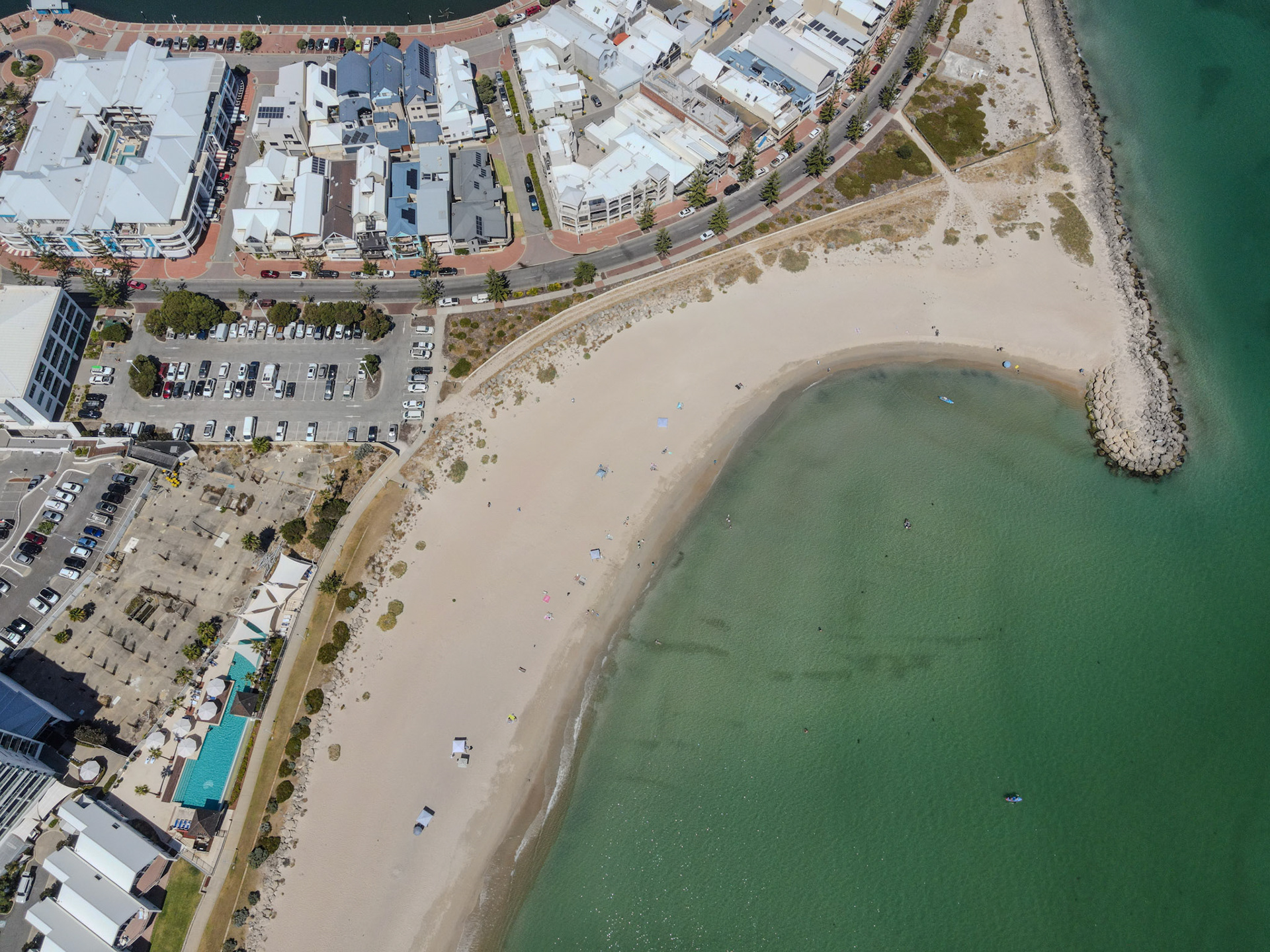 Overhead drone view of a curved sandy beach meeting calm green-blue water, with a rocky breakwater at one end. A beachfront promenade, parked cars and low-rise buildings border the sand; small umbrellas, swimmers and paddlecraft dot the shoreline and shallow bay. Pools, landscaped areas and a grid of streets and parking lots are visible behind the waterfront.