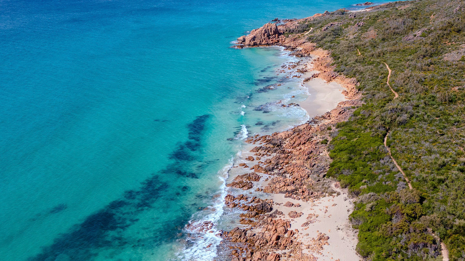 A stunning aerial view captures the pristine coastline, featuring turquoise waters gently lapping against rugged rocky cliffs. The vibrant landscape includes lush greenery bordering the beach, creating a harmonious blend of natural beauty. The clear sky enhances the serene and untouched environment, making it an ideal portrayal of nature's tranquility.
