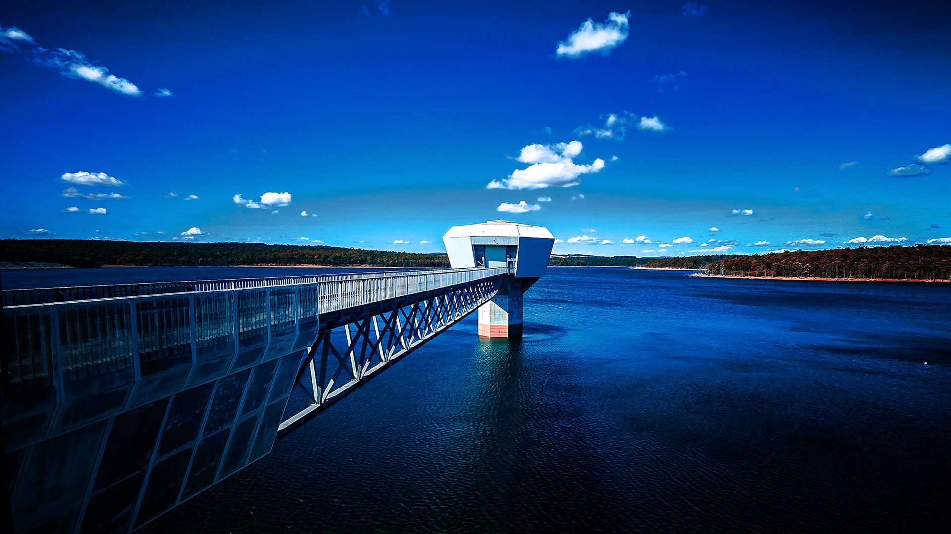 A long metal walkway stretches over deep blue water to a geometric intake tower perched on a single pillar. The calm reservoir is bordered by a dark tree line, while a vivid sky with scattered white clouds enhances the scene’s strong leading lines and crisp reflections.