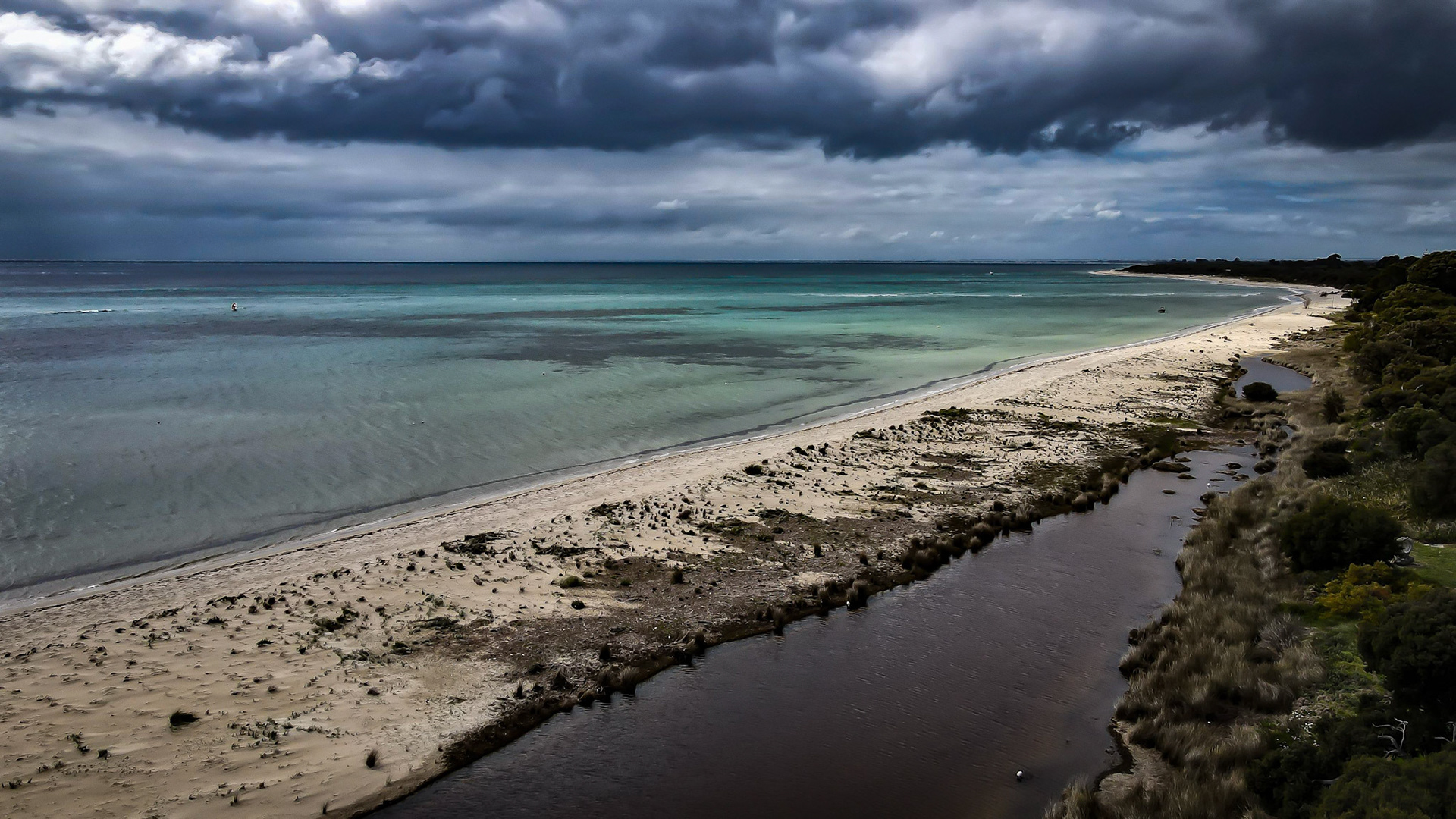 Captivating aerial view of a serene beach with a dramatic, overcast sky. The turquoise waters meet the sandy shoreline, with a dark cloudy sky creating a striking contrast. Lush greenery lines the riverbank, adding depth to the scenic panorama.