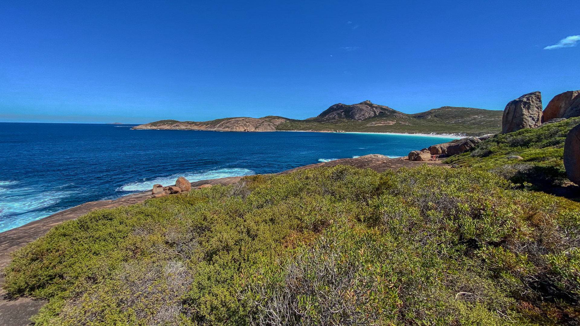 A scenic coastal landscape features a rugged coastline, deep blue ocean, and lush vegetation. Waves gently crash on the rocky shore.