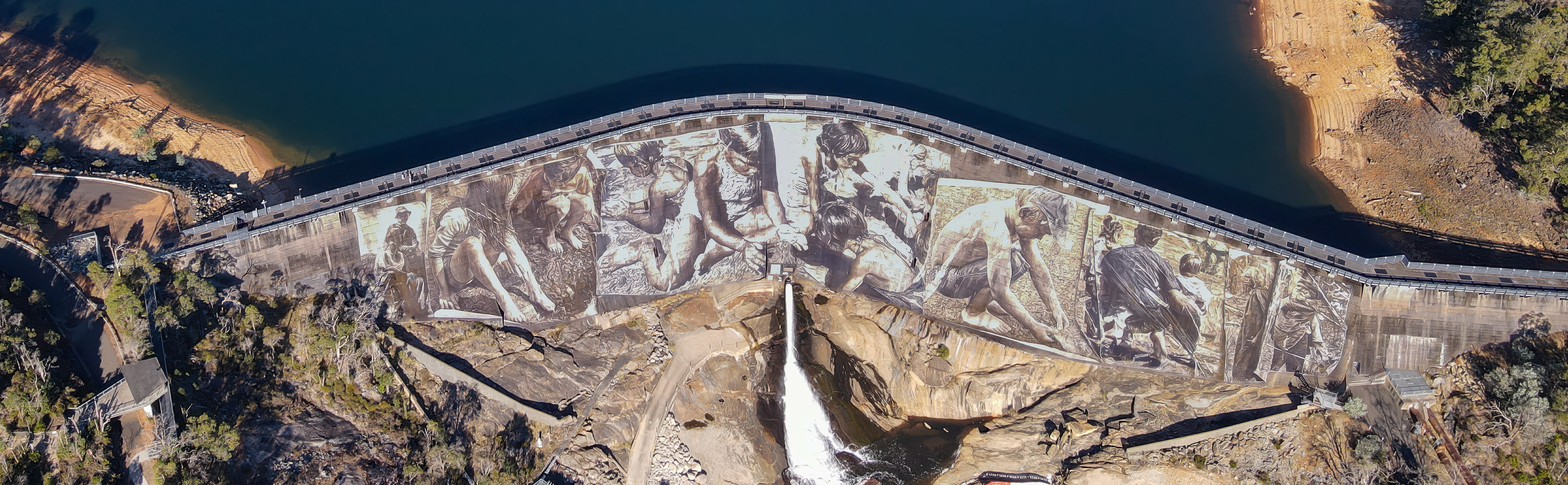 A high, curved concrete dam seen from above, with a vast sepia-toned mural stretching across its face. The artwork depicts people working and gathering in large, detailed scenes. Calm blue water fills the reservoir behind the wall, while a narrow spillway releases a white stream into a rocky gorge below. Surrounding hillsides are dotted with trees and scrub, with access roads and walkways tracing the edges of the structure.