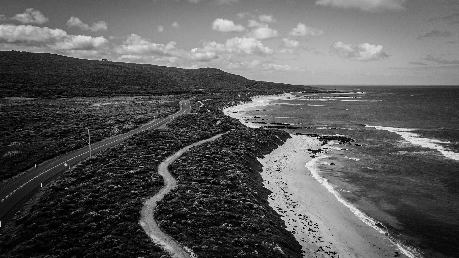 Aerial view of a winding coastal road running parallel to a pristine beach, captured in dramatic black and white. The image showcases the meandering pathway leading through lush vegetation along the rugged coastline, with gentle waves lapping the shore. Expansive ocean views highlight the natural beauty and tranquil atmosphere of this picturesque landscape under a sky dotted with soft clouds. The monochrome treatment adds a timeless and artistic quality to the scene, emphasizing contrasts and textures.
