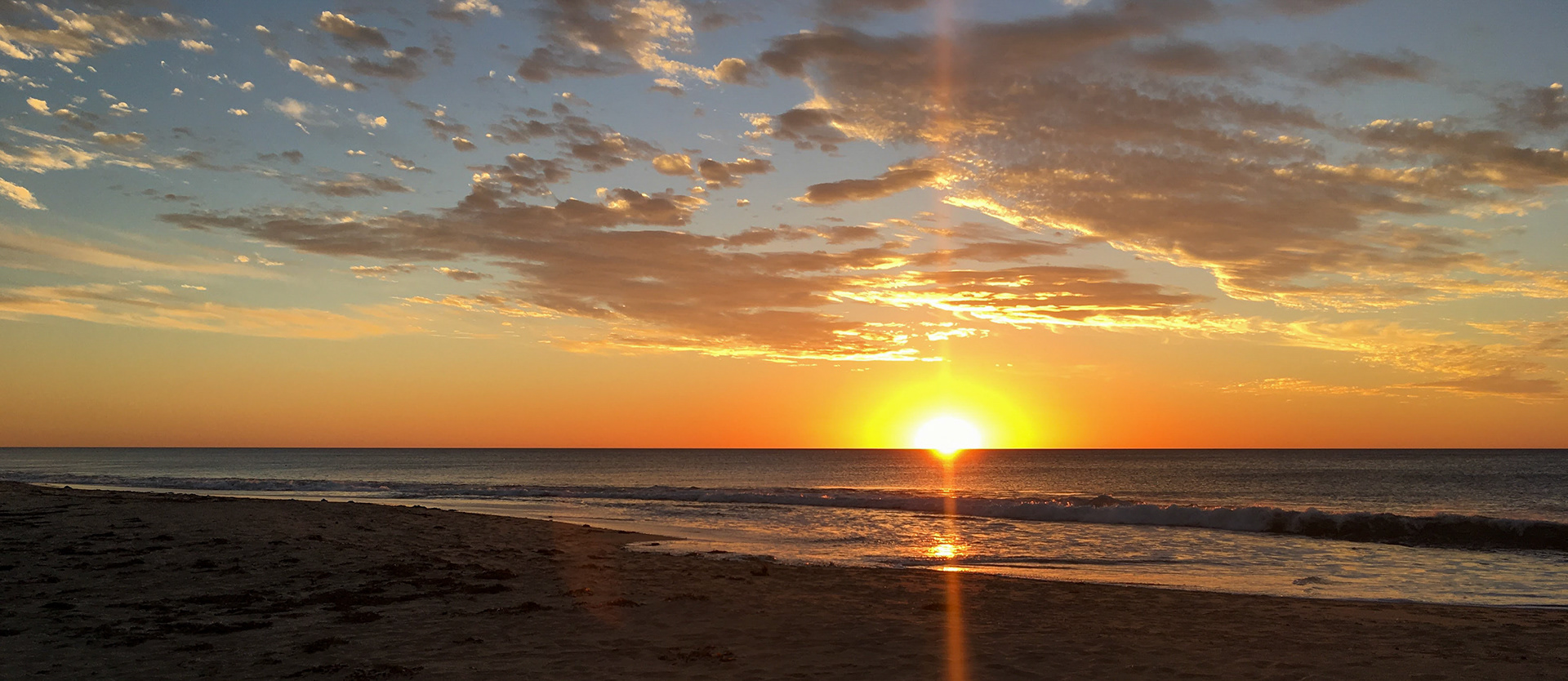 A serene sunset showcases warm hues of orange and yellow over the ocean, reflecting on gentle waves at the beach.