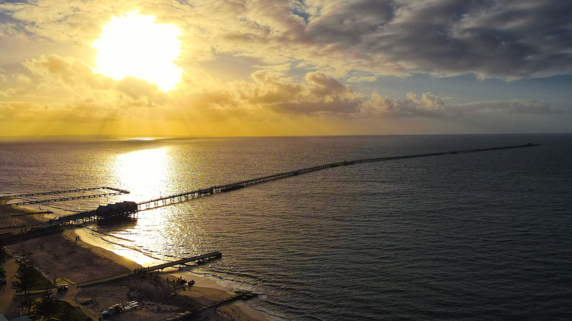 A breathtaking aerial view of a long pier extending into the ocean under a dramatic sunset. The golden sun casts its warm glow across the calm waters, creating a serene and picturesque landscape. Clouds hover above, adding depth to the sky, while the shoreline gently curves along the bottom of the image. Ideal for travel and nature themes.