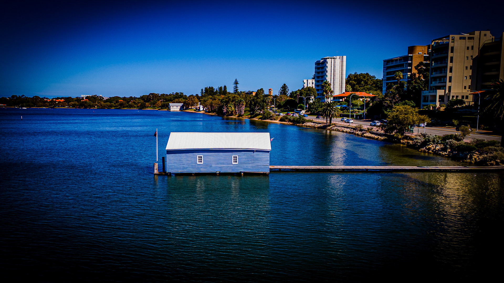 A delightful blue boathouse floats on calm waters, surrounded by lush trees and modern buildings. Sunlight sparkles on the surface, creating a peaceful midday scene.